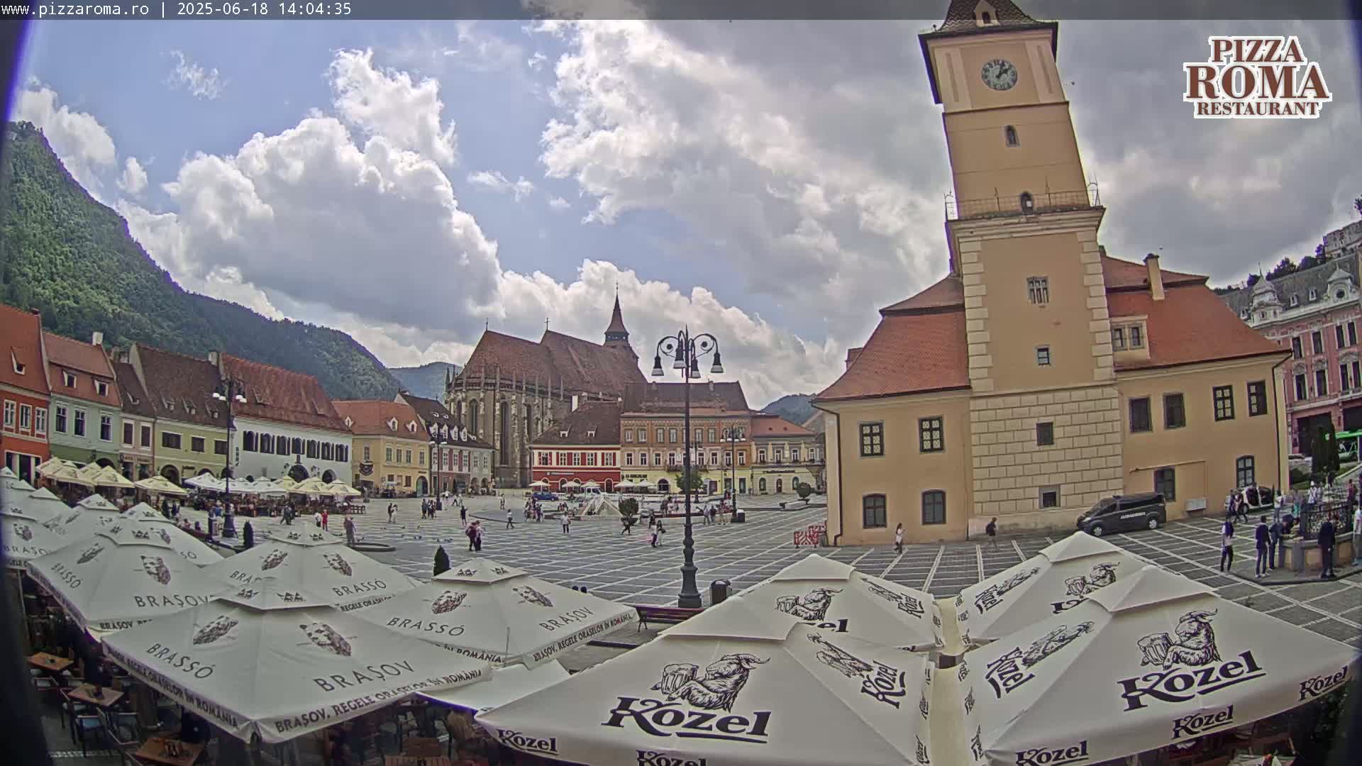 A town square with several buildings, including a tall clock tower, is partially shaded by many umbrellas under a partly cloudy sky.