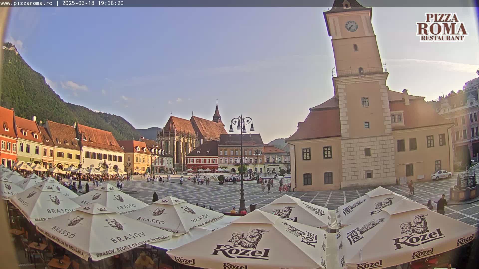A town square with several buildings, including a tall clock tower and a church, is filled with outdoor seating under umbrellas on a sunny day.