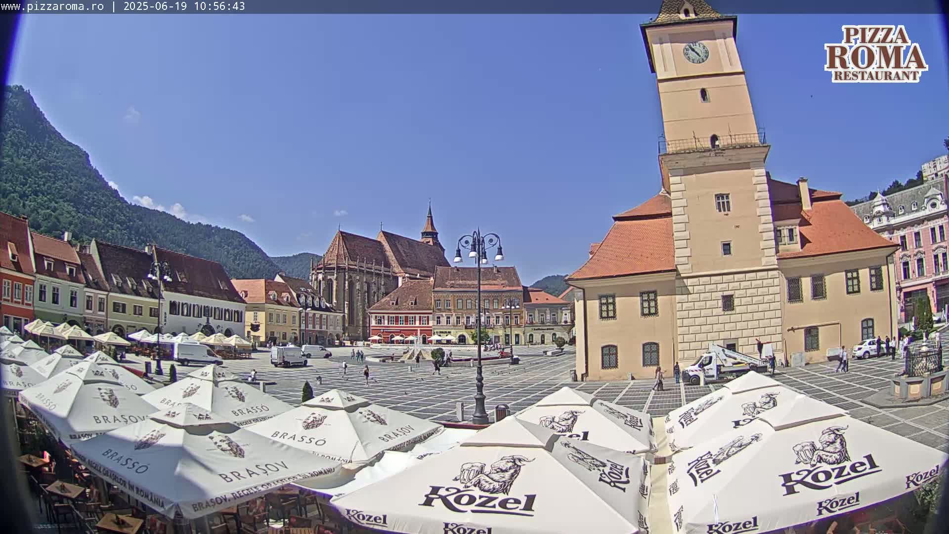 A sunny day in a European town square, with several outdoor café umbrellas and historic buildings surrounding a paved plaza, and mountains in the background.