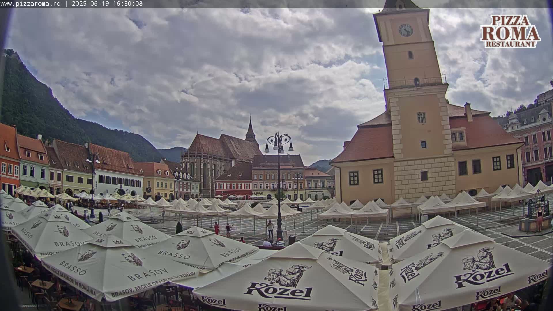 A town square filled with numerous market umbrellas, surrounded by historic buildings under a partly cloudy sky.
