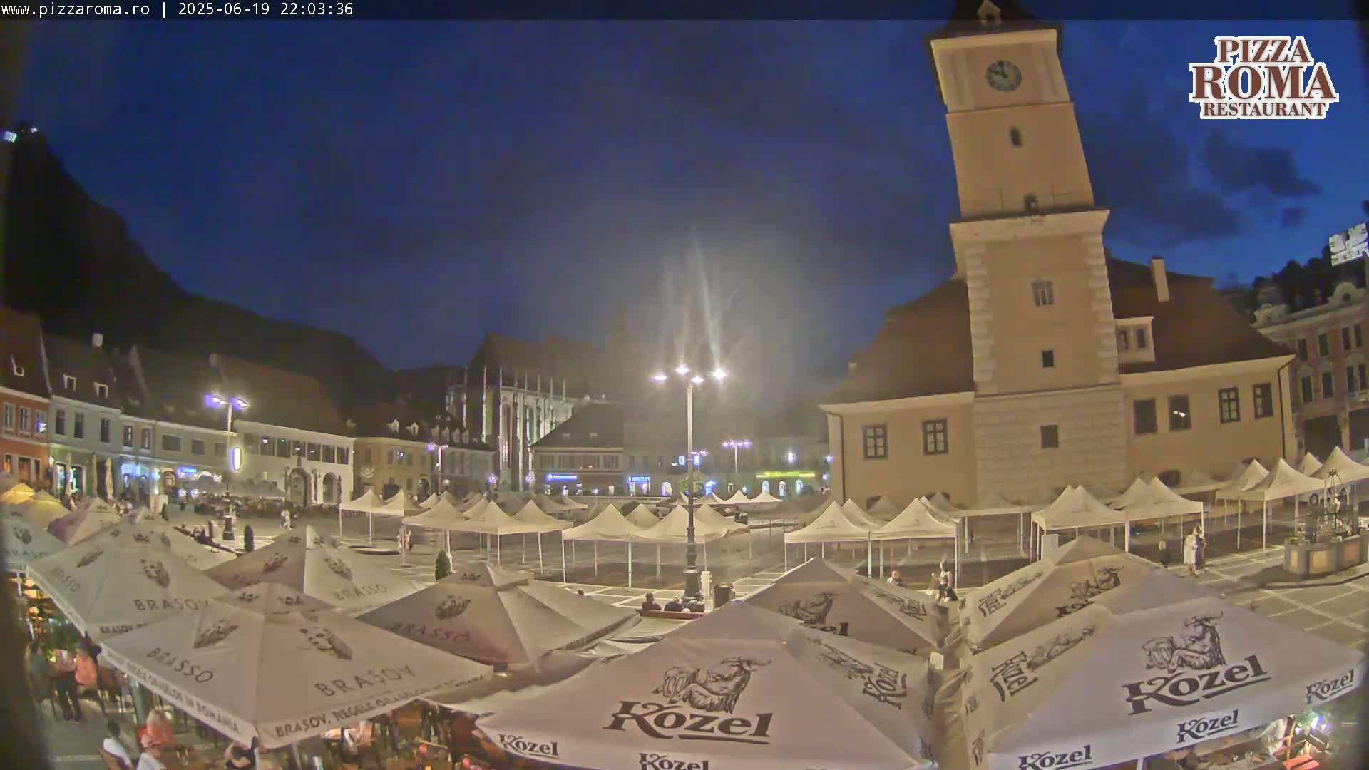 A nighttime view of a town square filled with numerous market umbrellas and a tall clock tower under a dark blue sky.