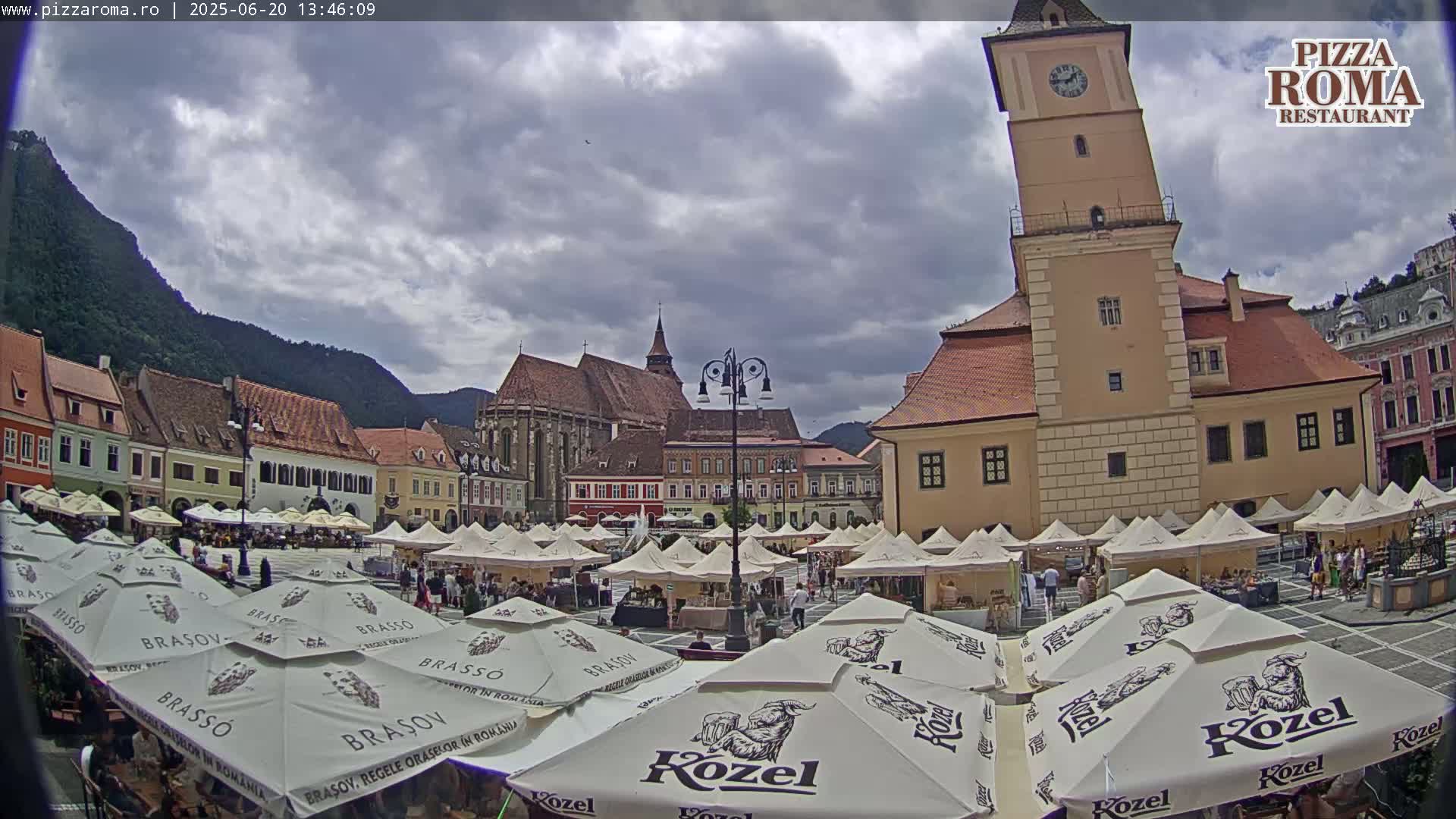 A town square, under a partly cloudy sky, features numerous market stalls and umbrellas, with a large, leaning clock tower and historic buildings in the background.