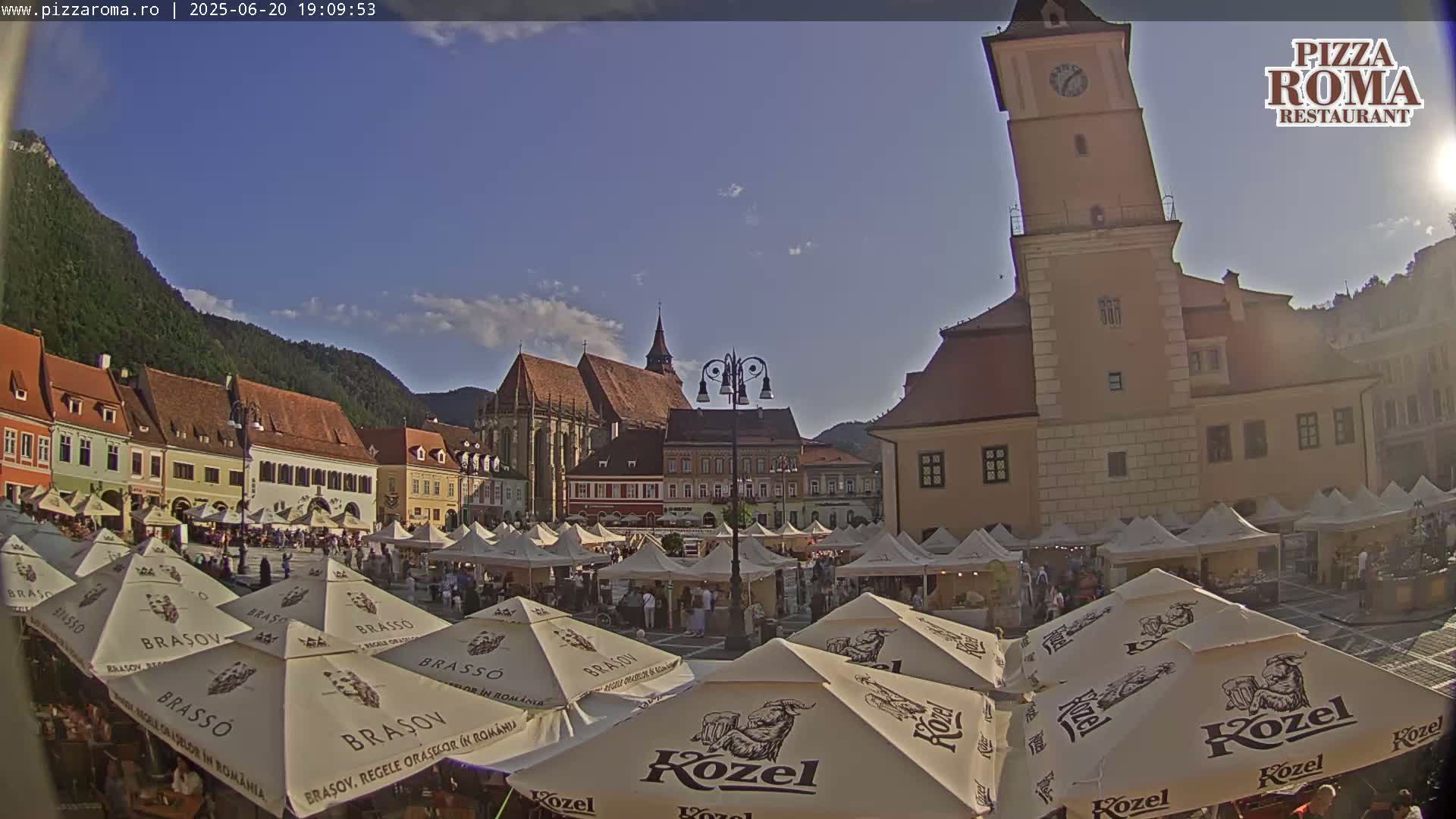 A sunny town square filled with market stalls under beige umbrellas, surrounded by historical buildings, including a tall clock tower, with mountains in the background.
