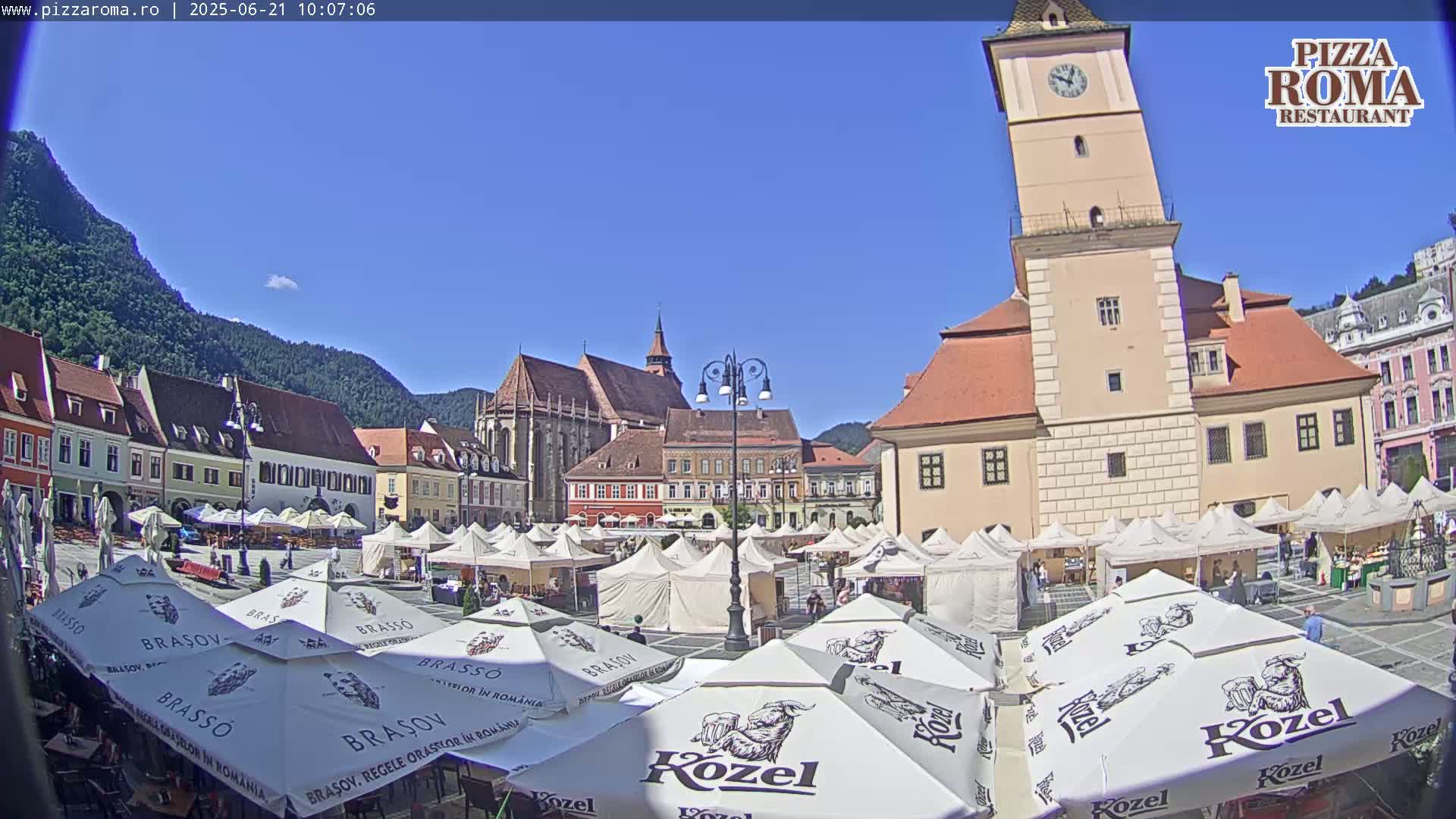 A sunny town square with numerous market stalls and outdoor seating under branded umbrellas, surrounded by historic buildings and mountains in the background.