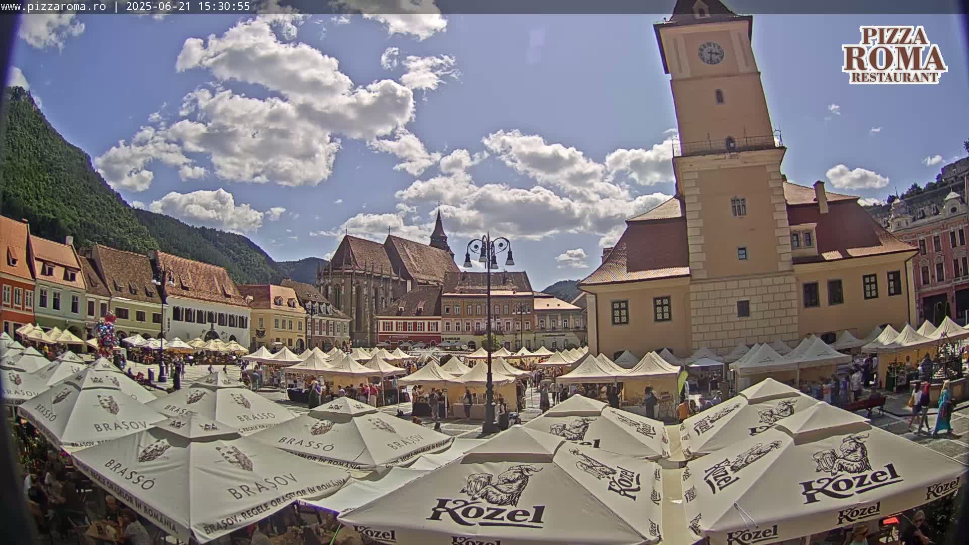 A sunny day in a European town square, filled with market stalls under numerous parasols, with a large, tall building and mountains in the background.
