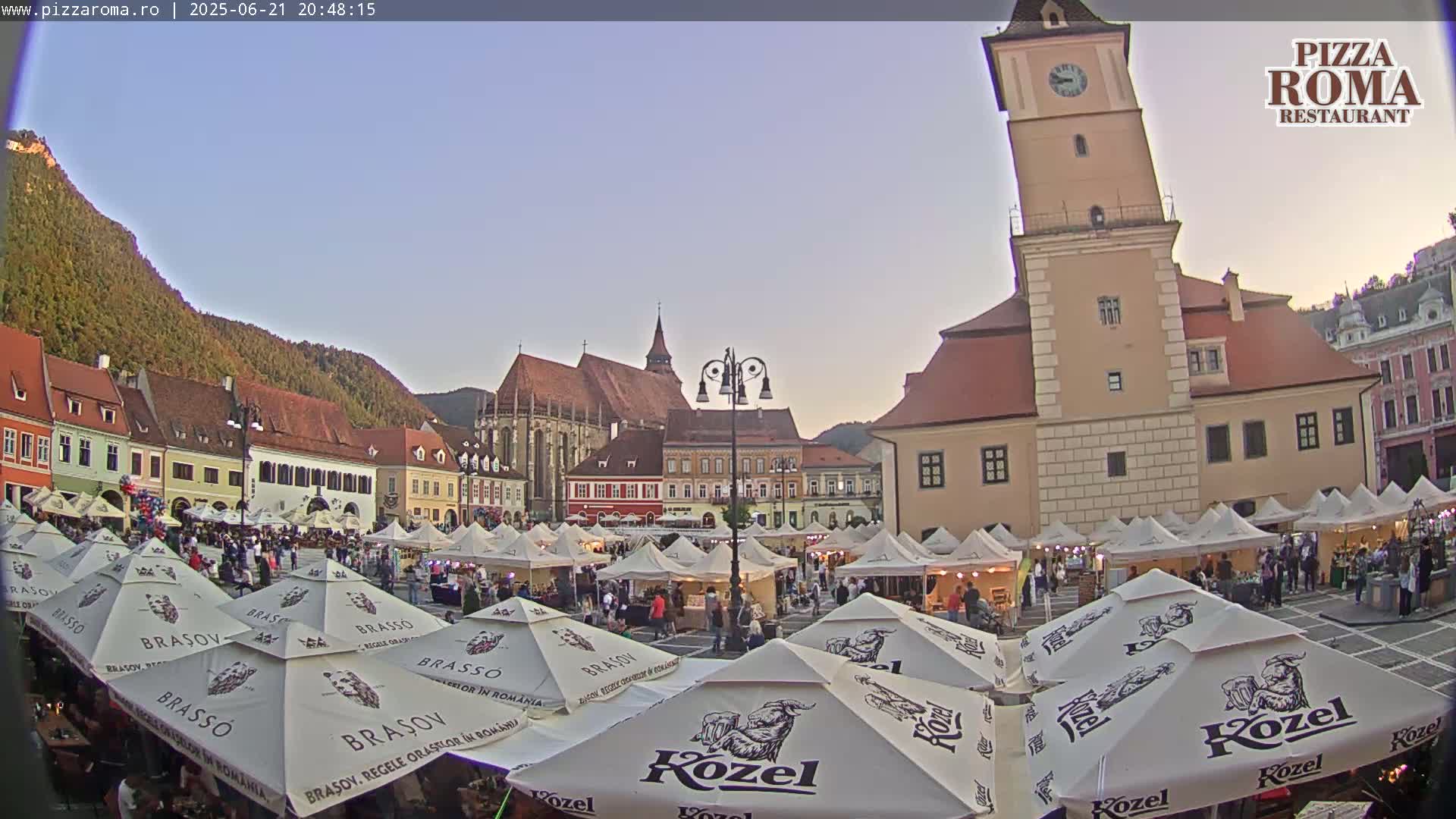 A town square filled with market stalls under a twilight sky, with a tall clock tower and older buildings visible in the background.