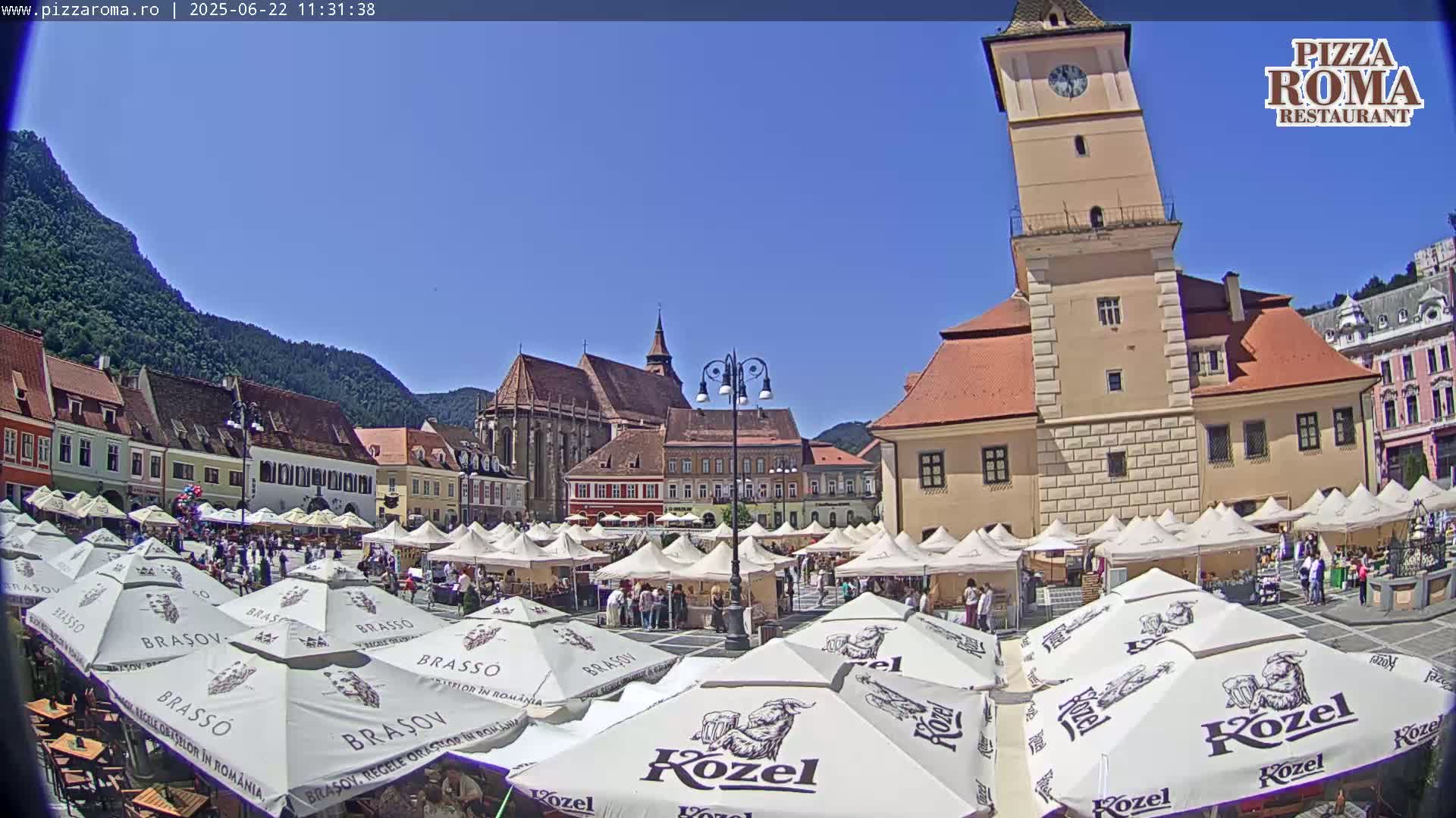 A sunny day in a European town square filled with market stalls and outdoor seating under parasols, with historic buildings and a tall clock tower in the background.