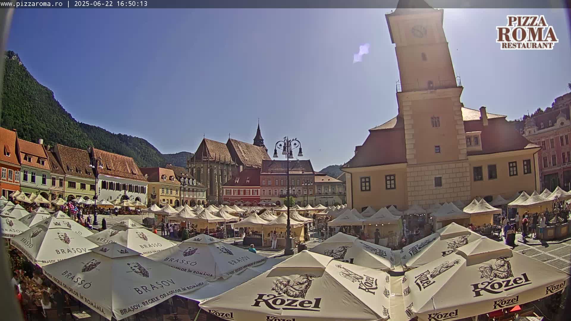 A sunny town square filled with market stalls and people under numerous parasols, with a tall clock tower and older buildings surrounding it, backed by mountains.