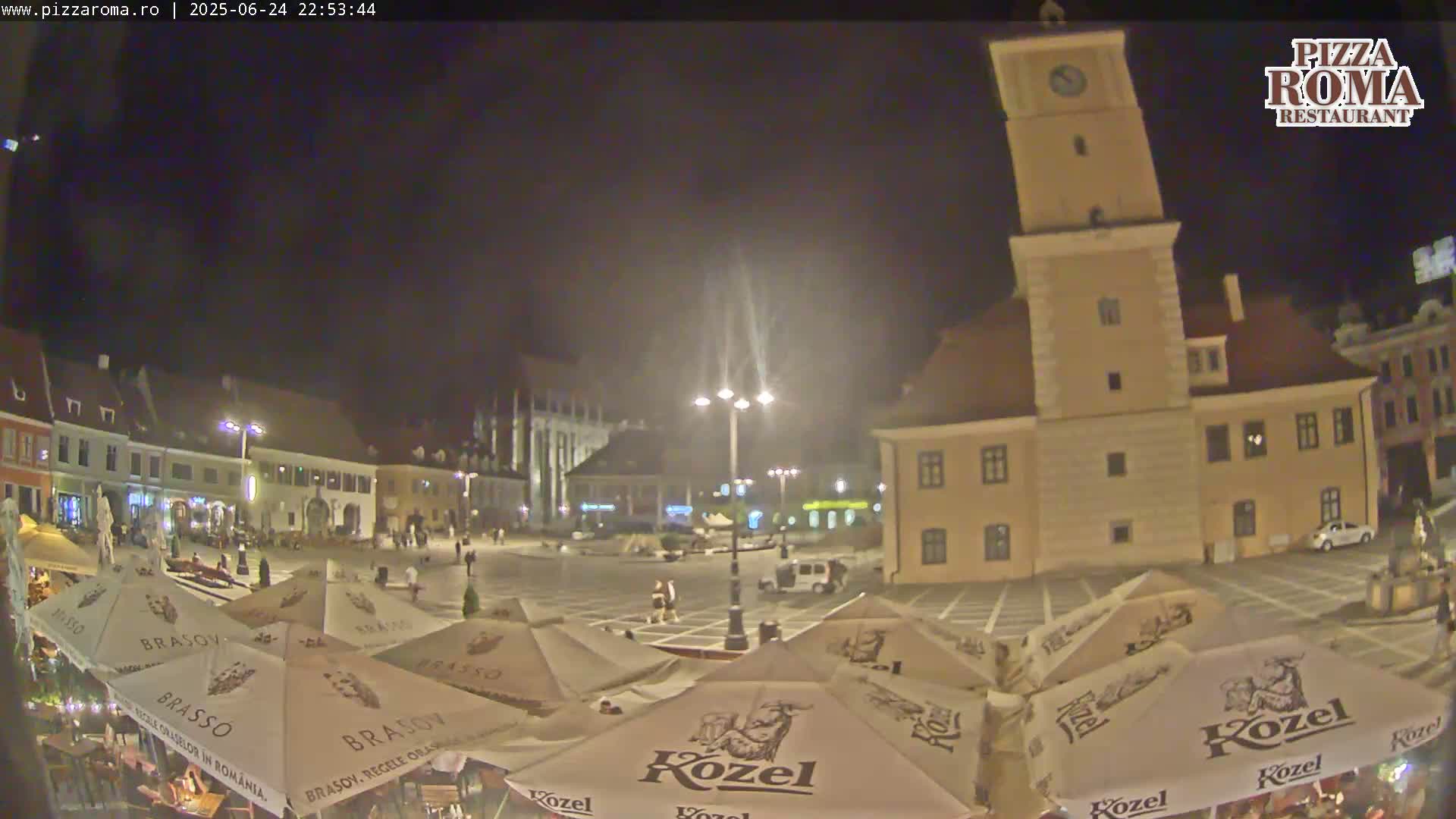 A nighttime view of a town square with several buildings, outdoor seating under umbrellas, and a tall clock tower.