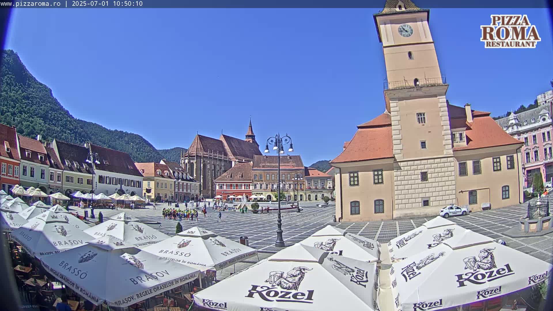 A sunny day in a European town square shows several buildings, including a tall clock tower, with outdoor cafe umbrellas in the foreground.