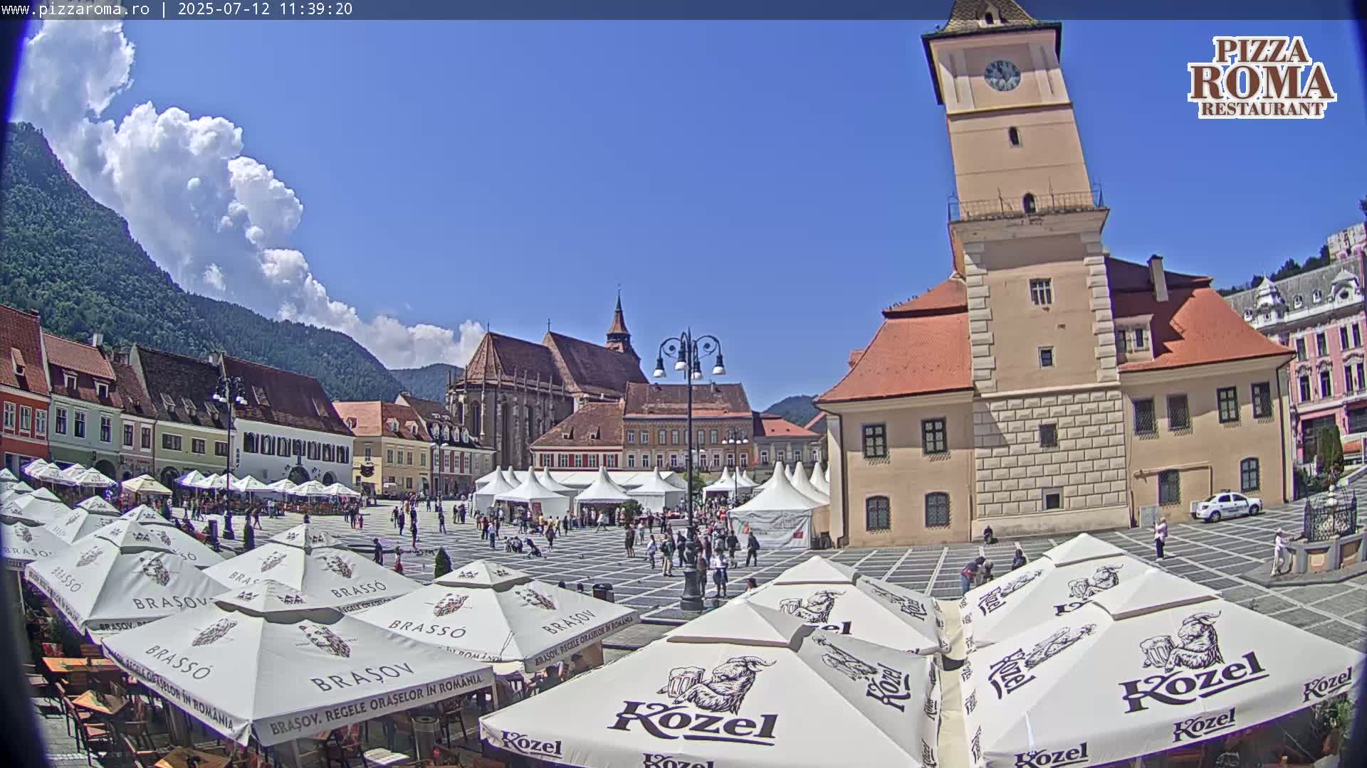 A sunny town square, surrounded by medieval-style buildings and mountains in the background, features numerous outdoor umbrellas and people under a mostly clear sky with some cumulus clouds.