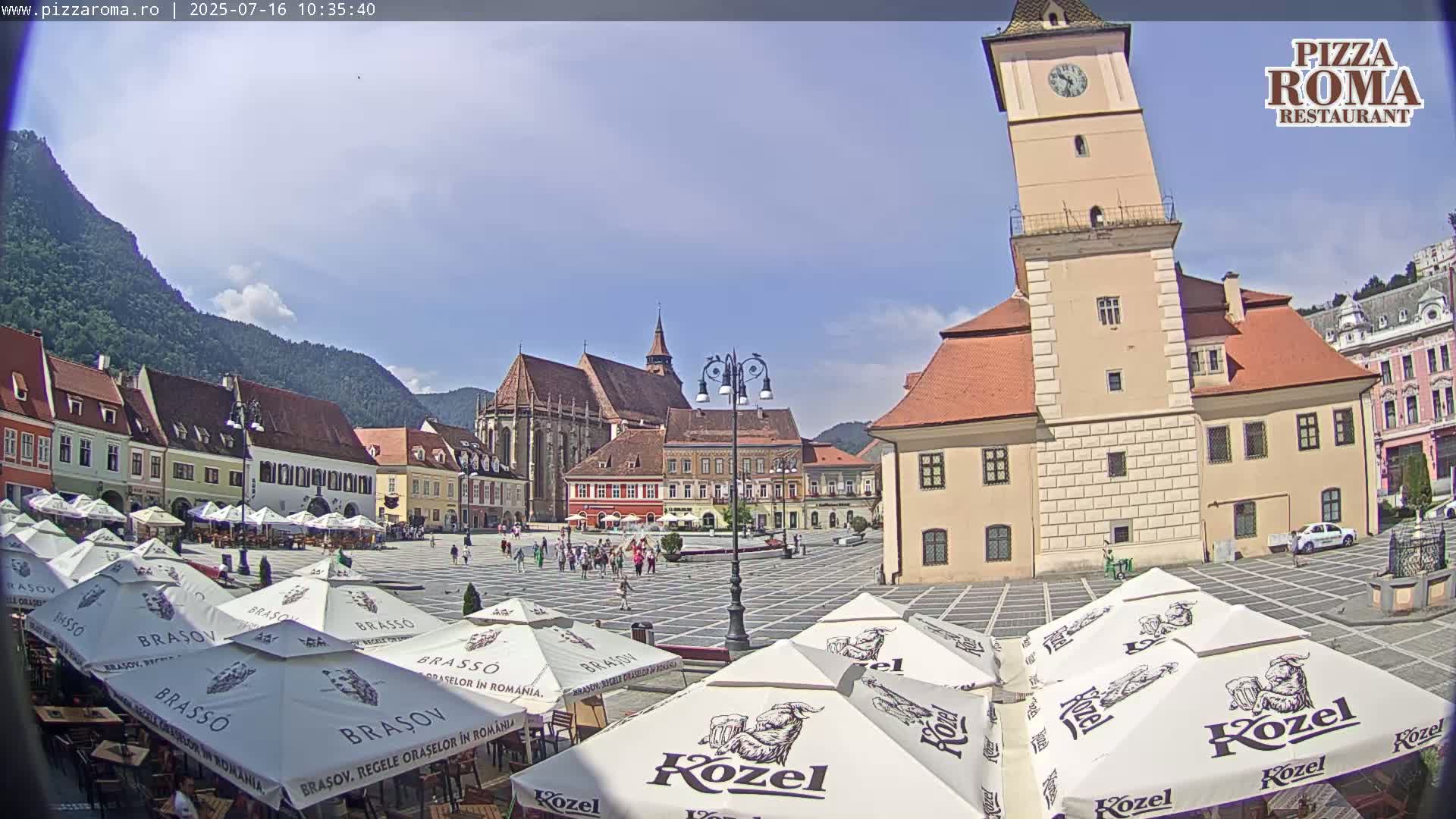 A sunny town square with several buildings, a leaning tower, and numerous outdoor cafe umbrellas.
