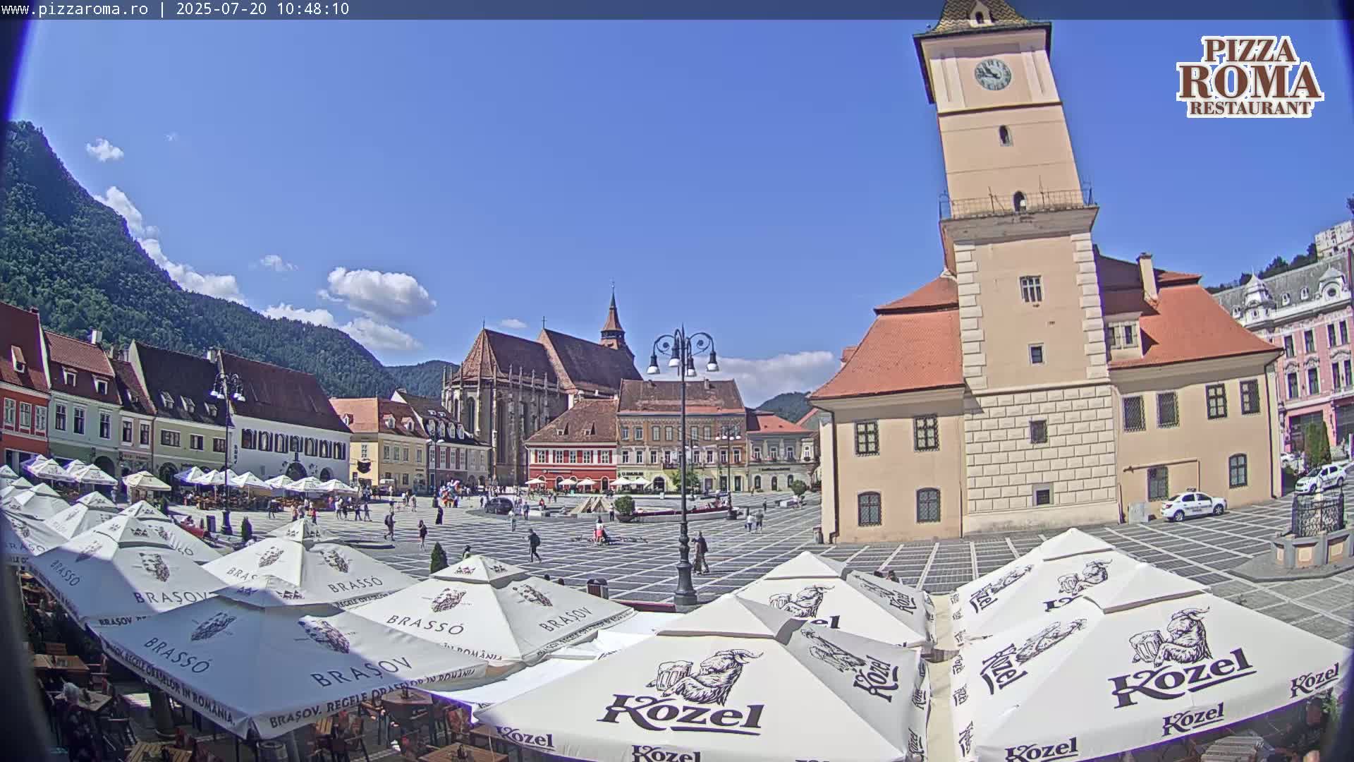 A sunny town square with several buildings, including a tall clock tower, is surrounded by outdoor seating with numerous umbrellas.