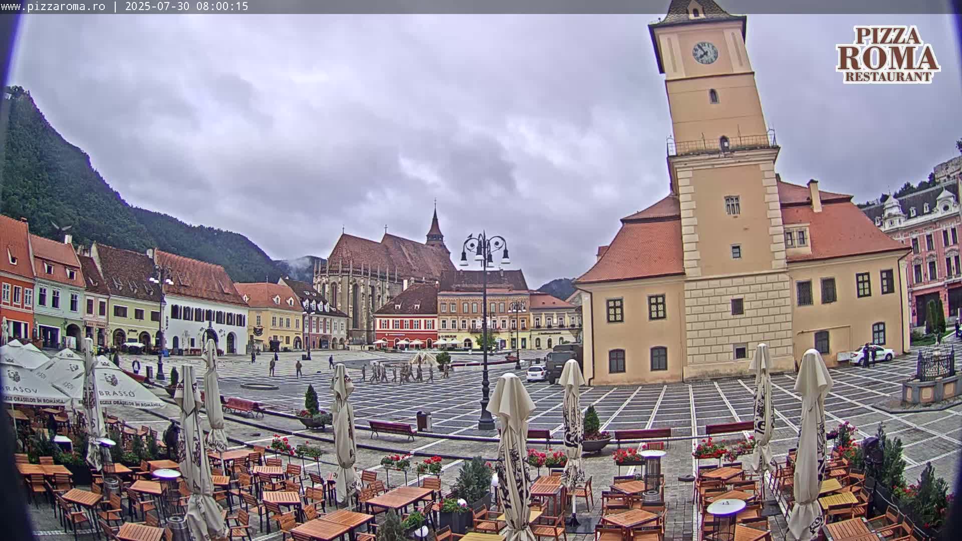 A sunny town square with several buildings, including a tall clock tower, is surrounded by outdoor seating with numerous umbrellas.