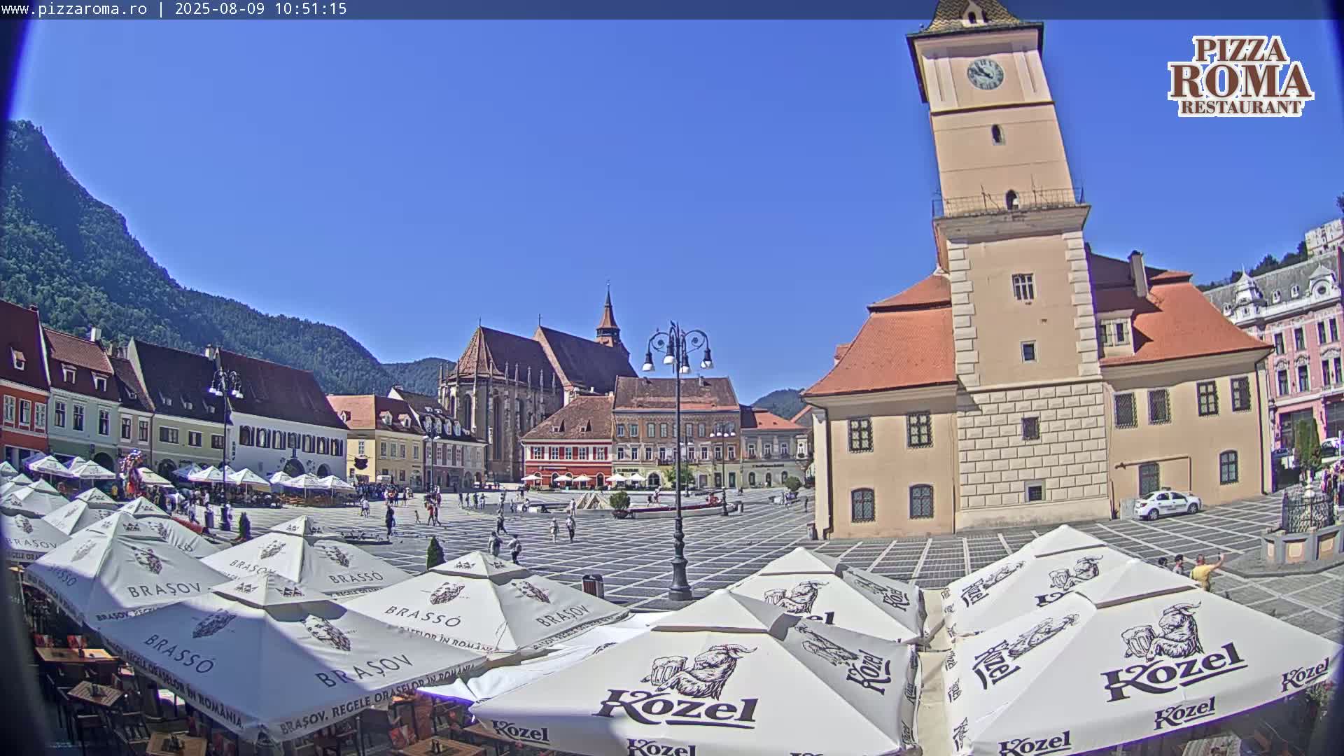 A town square with several buildings, a tall clock tower, and numerous outdoor cafe umbrellas under a clear blue sky.