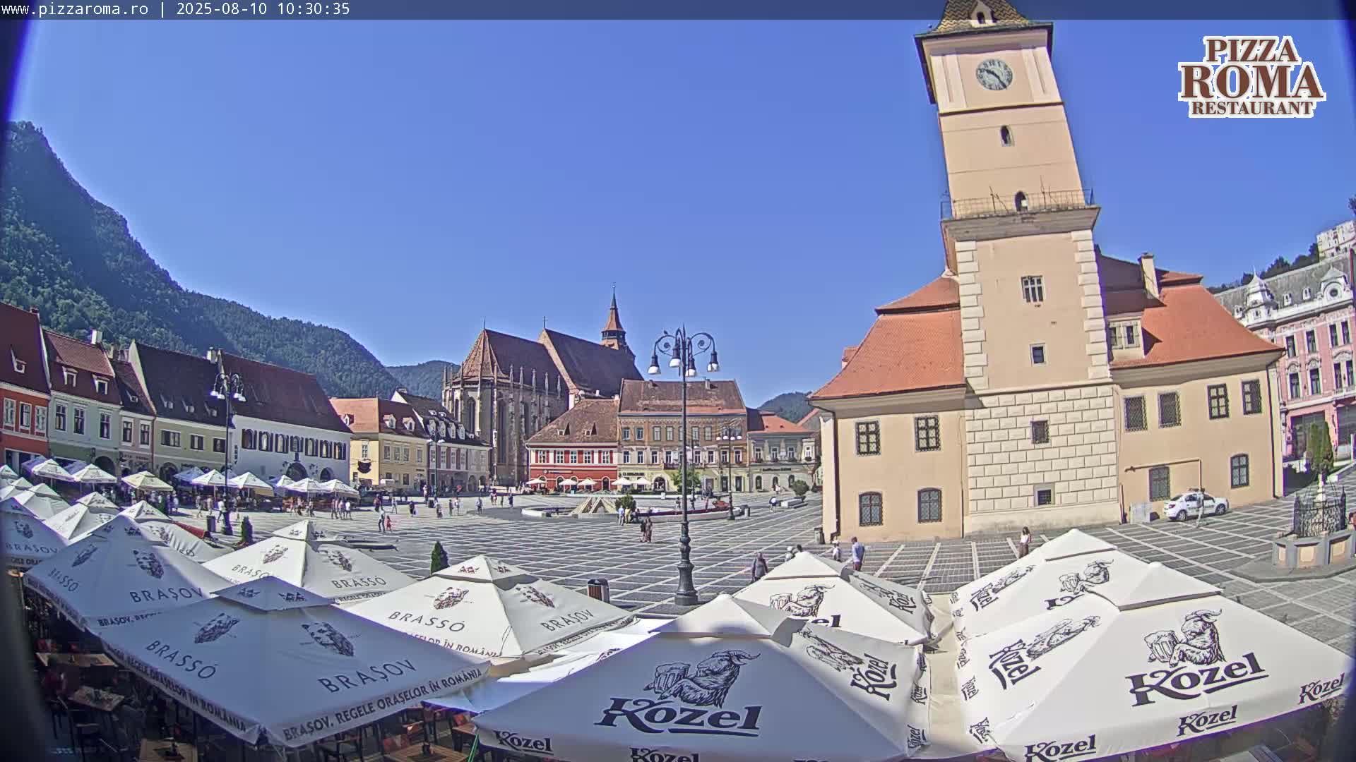 A town square with numerous outdoor cafe umbrellas, historic buildings, and a tall clock tower is visible under a clear blue sky.