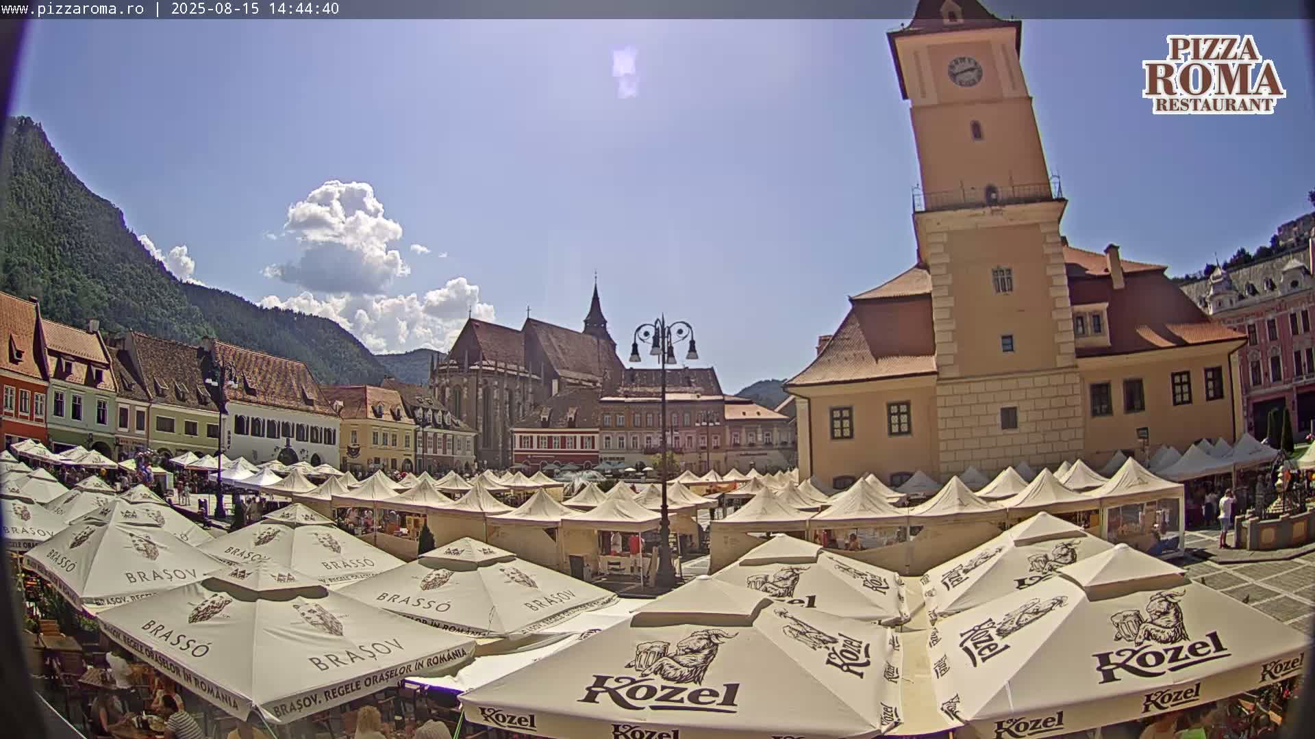 A town square with numerous outdoor cafe umbrellas, historic buildings, and a tall clock tower is visible under a clear blue sky.