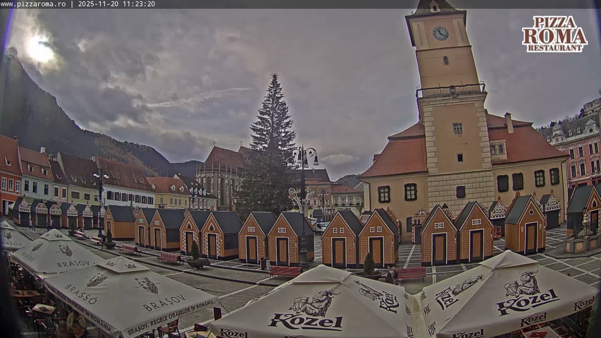 Under an overcast sky, a European town square is seen featuring a tall Christmas tree, a prominent clock tower, a large church, and rows of small, decorative market stalls, with distant mountains completing the scene.