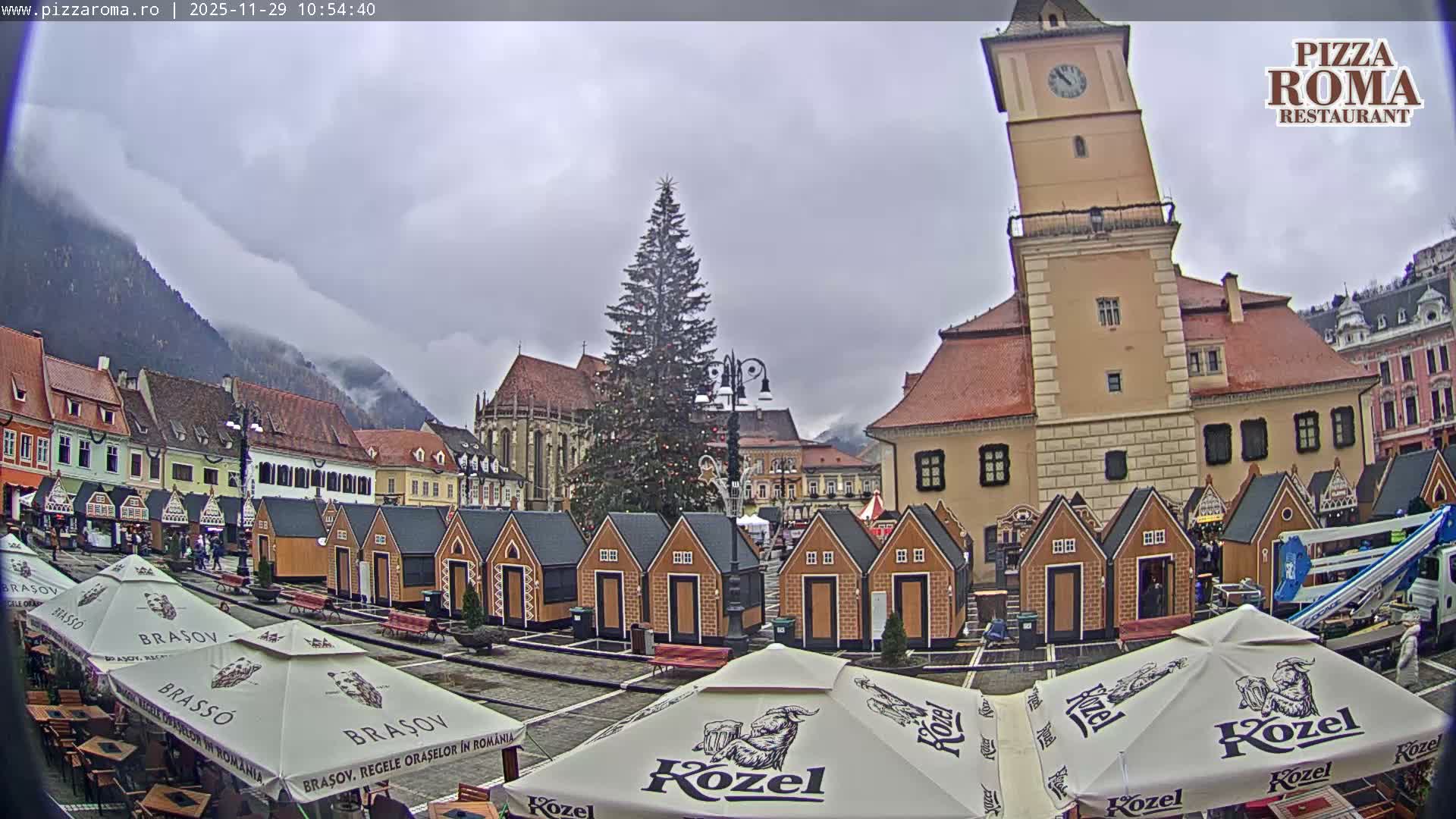 An outdoor European town square on an overcast day features a large decorated Christmas tree, numerous festive market stalls, historic architecture, and distant misty mountains.