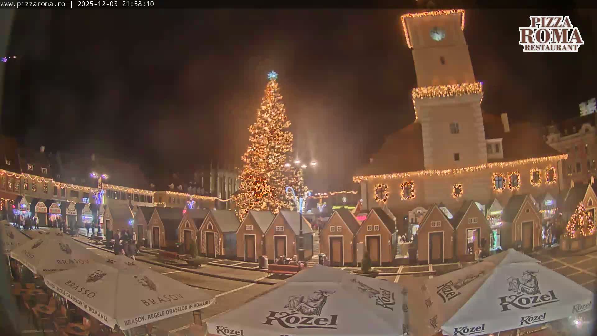 A bustling Christmas market square is illuminated at night with a large decorated tree, festive lights on buildings including a clock tower, rows of small stalls, and people strolling amidst outdoor umbrellas on a clear night.