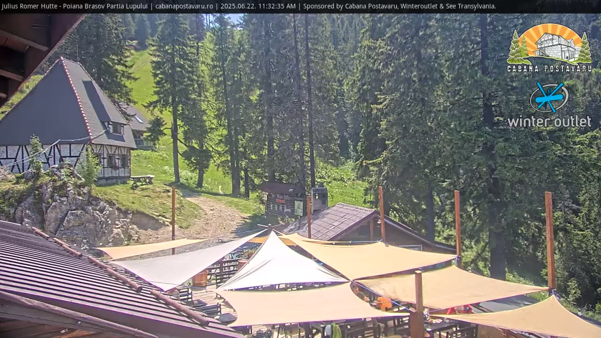 A sunny outdoor scene shows a mountainside with several buildings, including a rustic-style house and a patio area covered by tan shade sails.