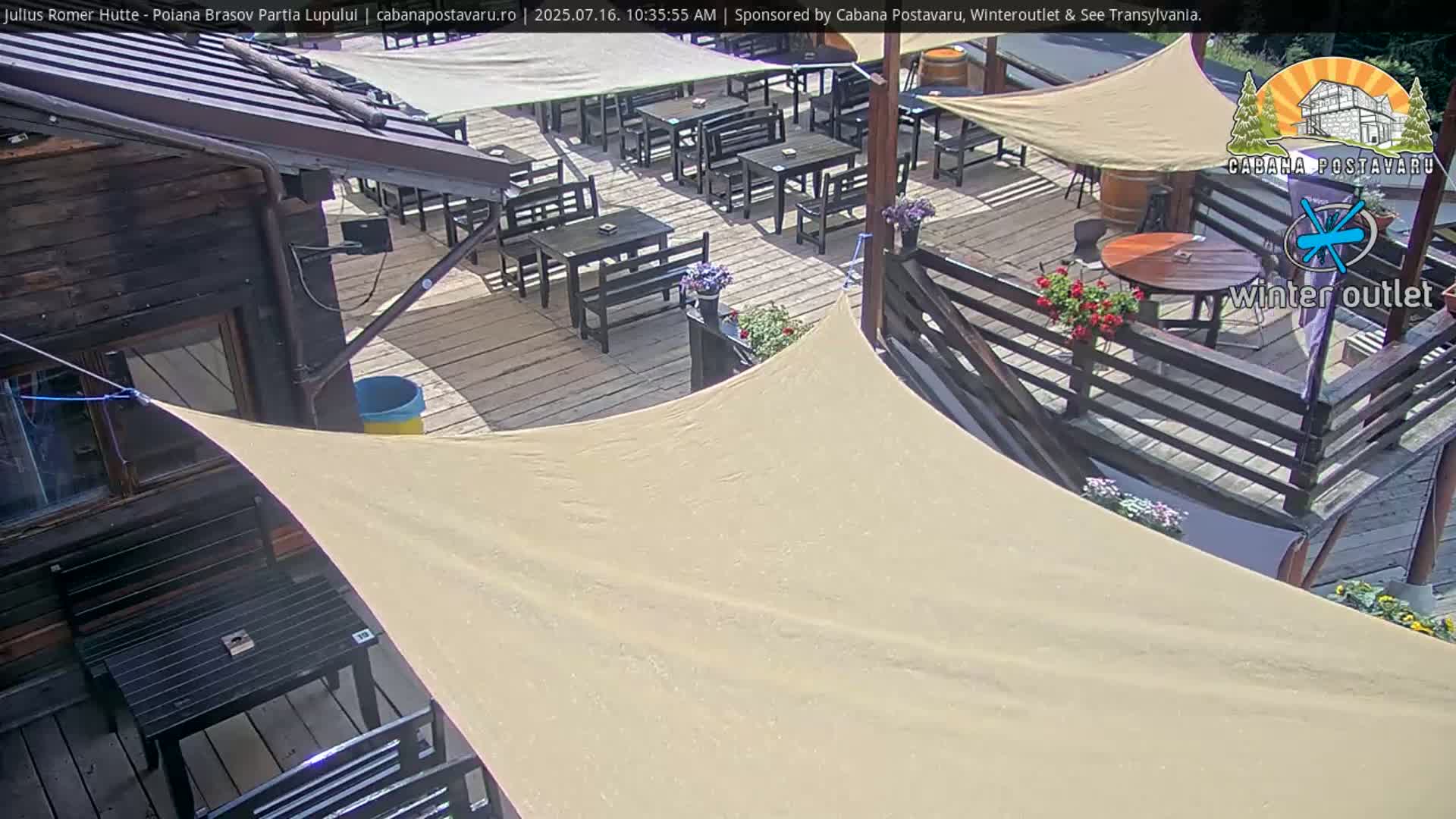 A sunny outdoor patio scene with numerous tables and chairs set up on a wooden deck, partially shaded by beige canopies, adjacent to a wooden building.