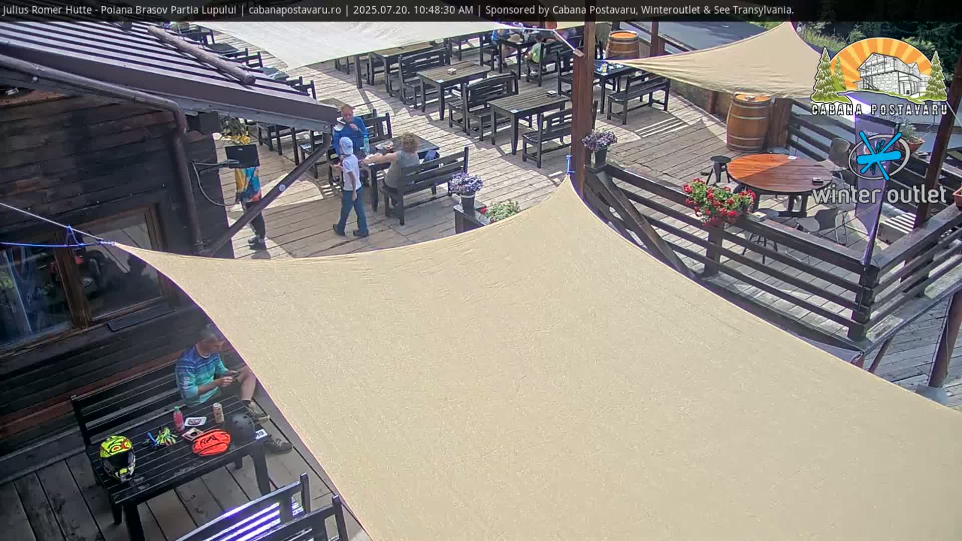 A sunny outdoor patio area with numerous tables and chairs, a wooden cabin, and people sitting at tables under a beige shade structure.