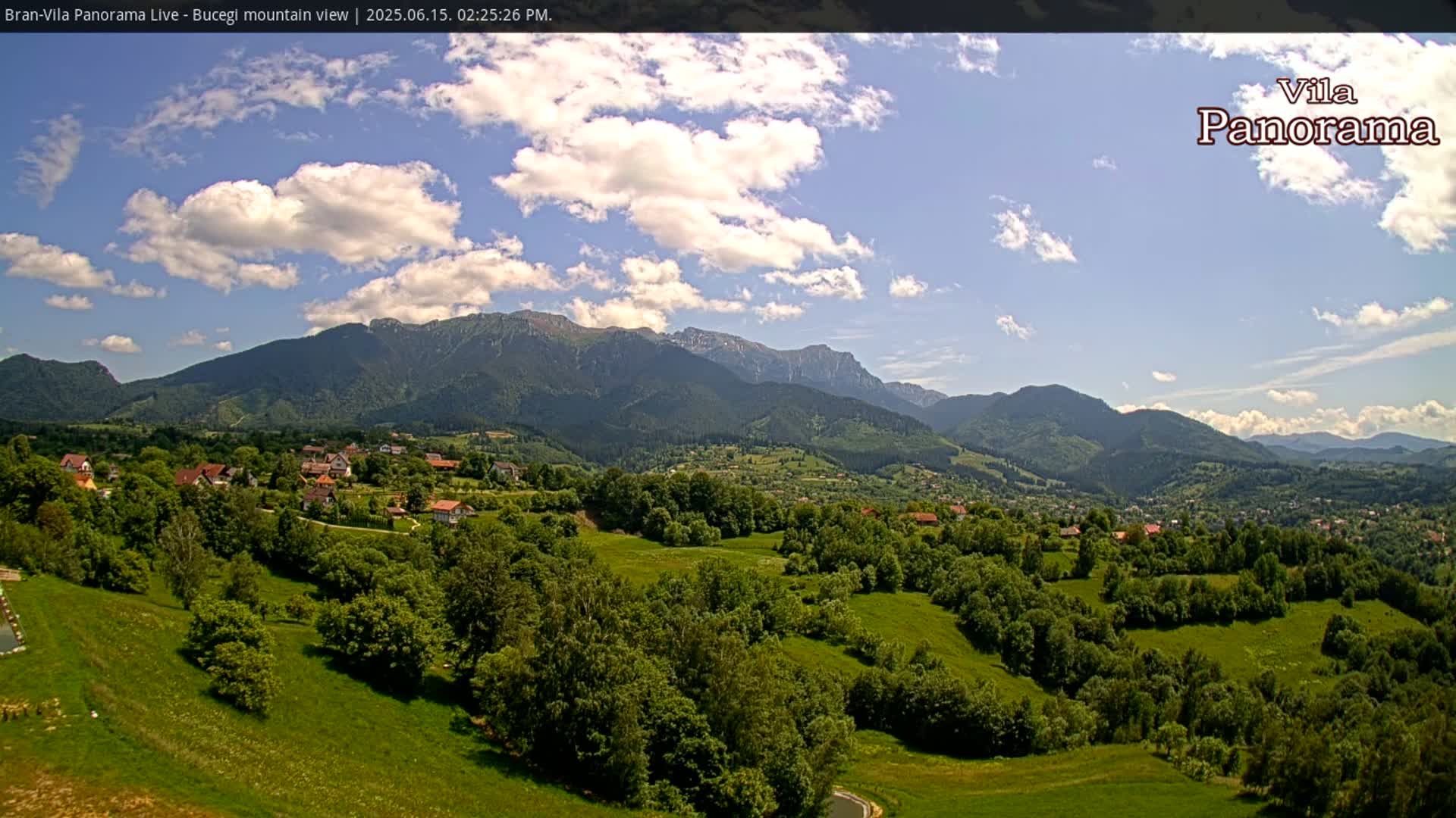 A partly cloudy sky overlooks a verdant valley dotted with houses and trees, nestled against a backdrop of imposing mountains.