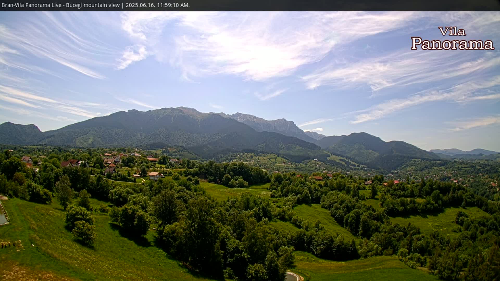 A sprawling green valley dotted with houses and trees, nestled beneath a range of mountains under a mostly clear, bright sky.