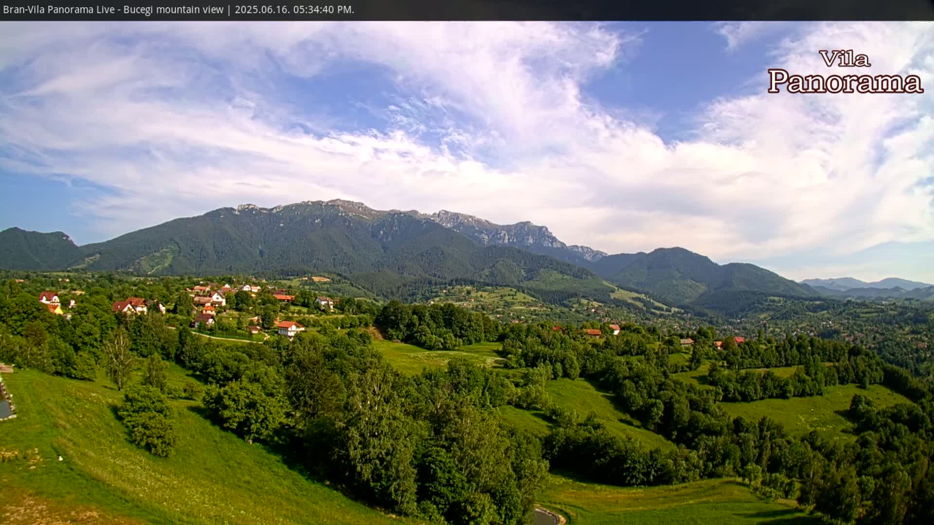 A partly cloudy sky overlooks a verdant valley dotted with houses and trees, leading to a range of forested mountains in the distance.