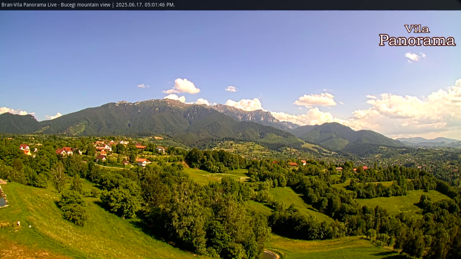 A picturesque village nestled in a verdant valley, with a range of mountains under a partly cloudy blue sky.