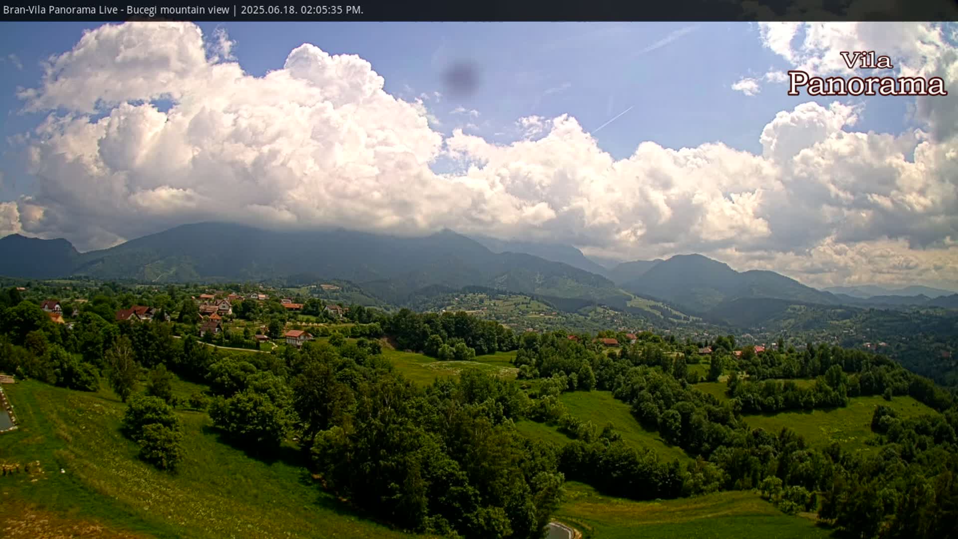 A partly cloudy sky hangs over a verdant valley dotted with houses and trees, nestled against a backdrop of rolling mountains.
