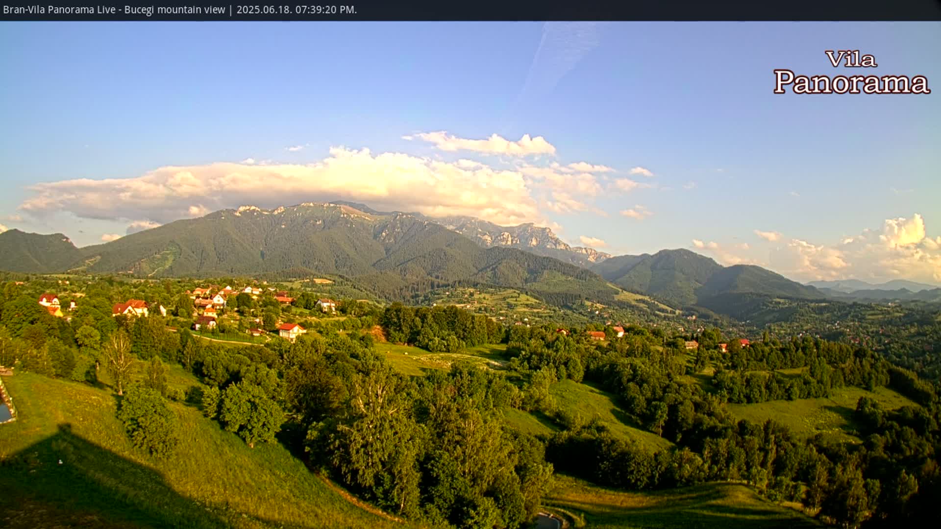 A partly cloudy evening sky overlooks a verdant valley of houses nestled amongst rolling green hills and a mountain range in the background.