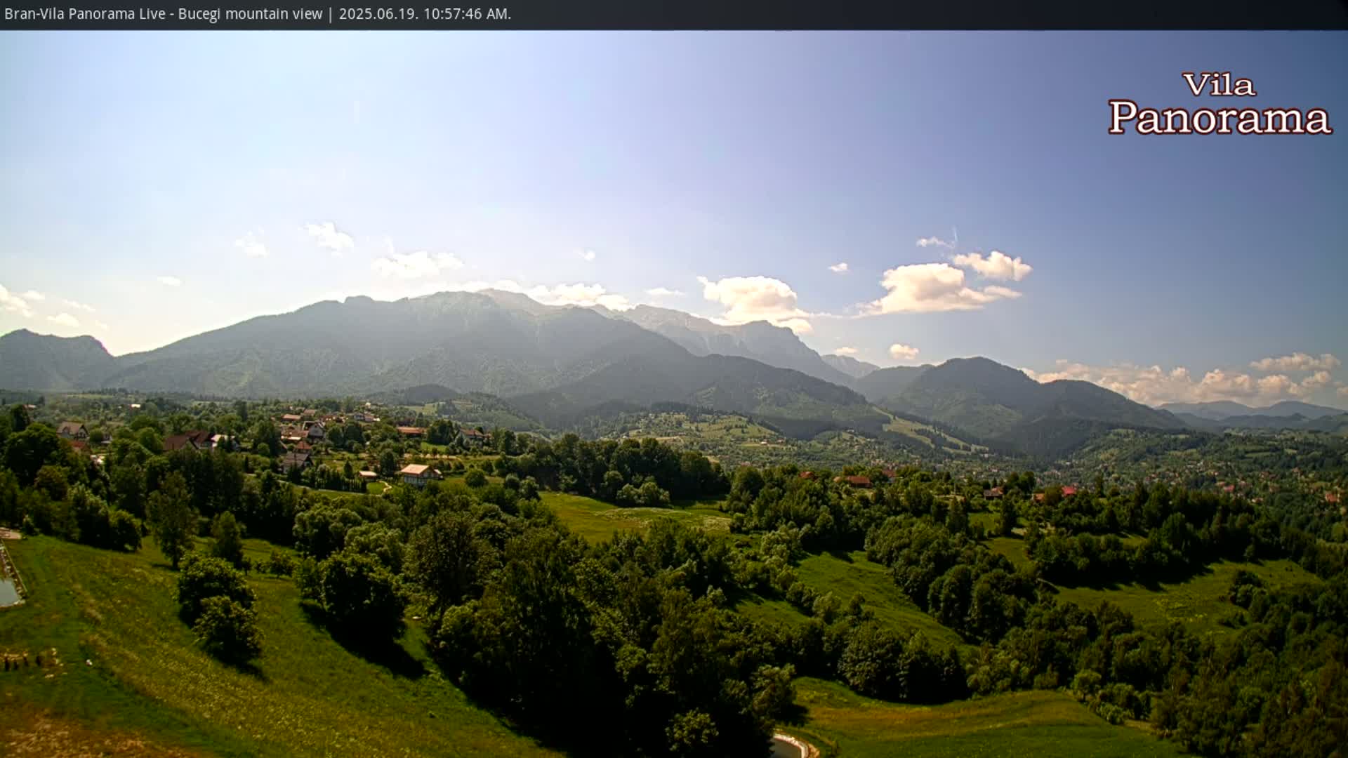 A sunny day reveals a picturesque landscape of rolling green hills dotted with houses, leading to a majestic mountain range under a partly cloudy sky.