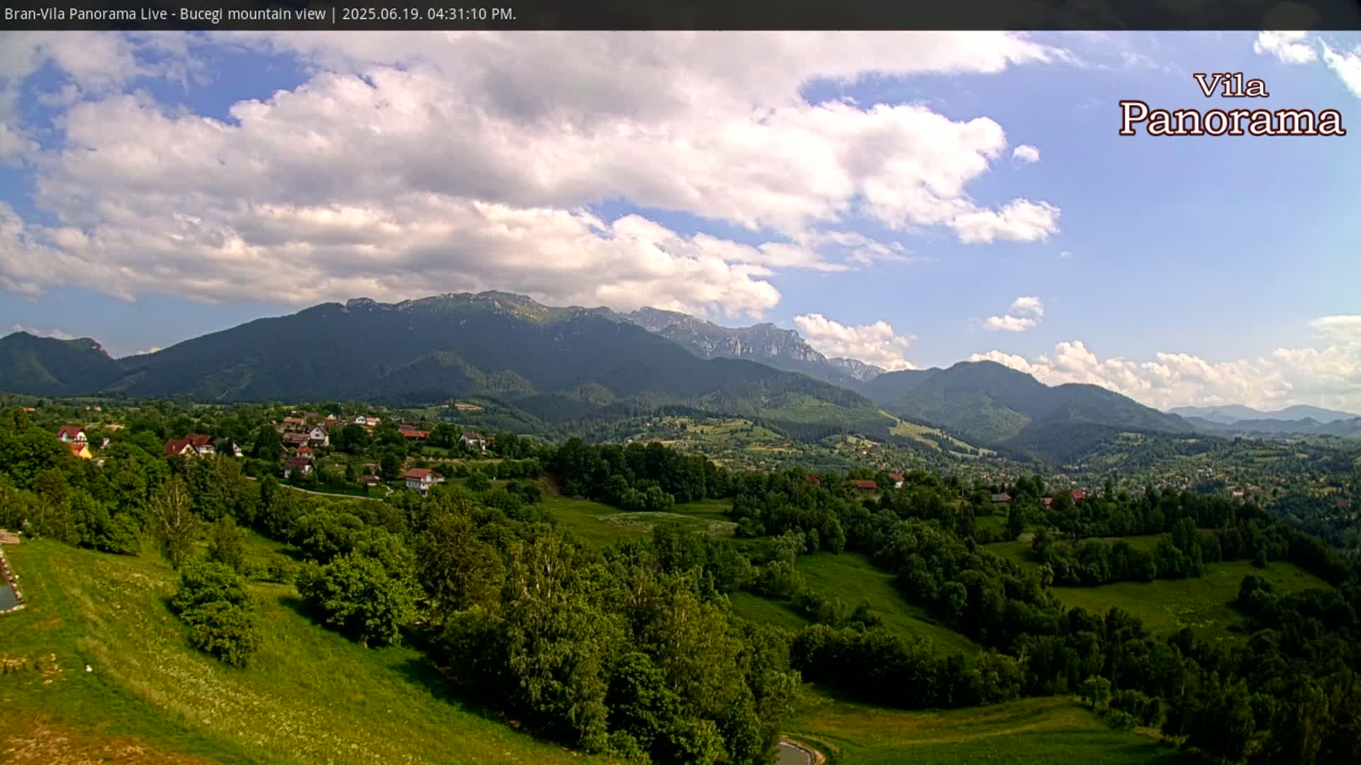 A partly cloudy daytime view of a village nestled in a valley, surrounded by lush green hills and mountains.