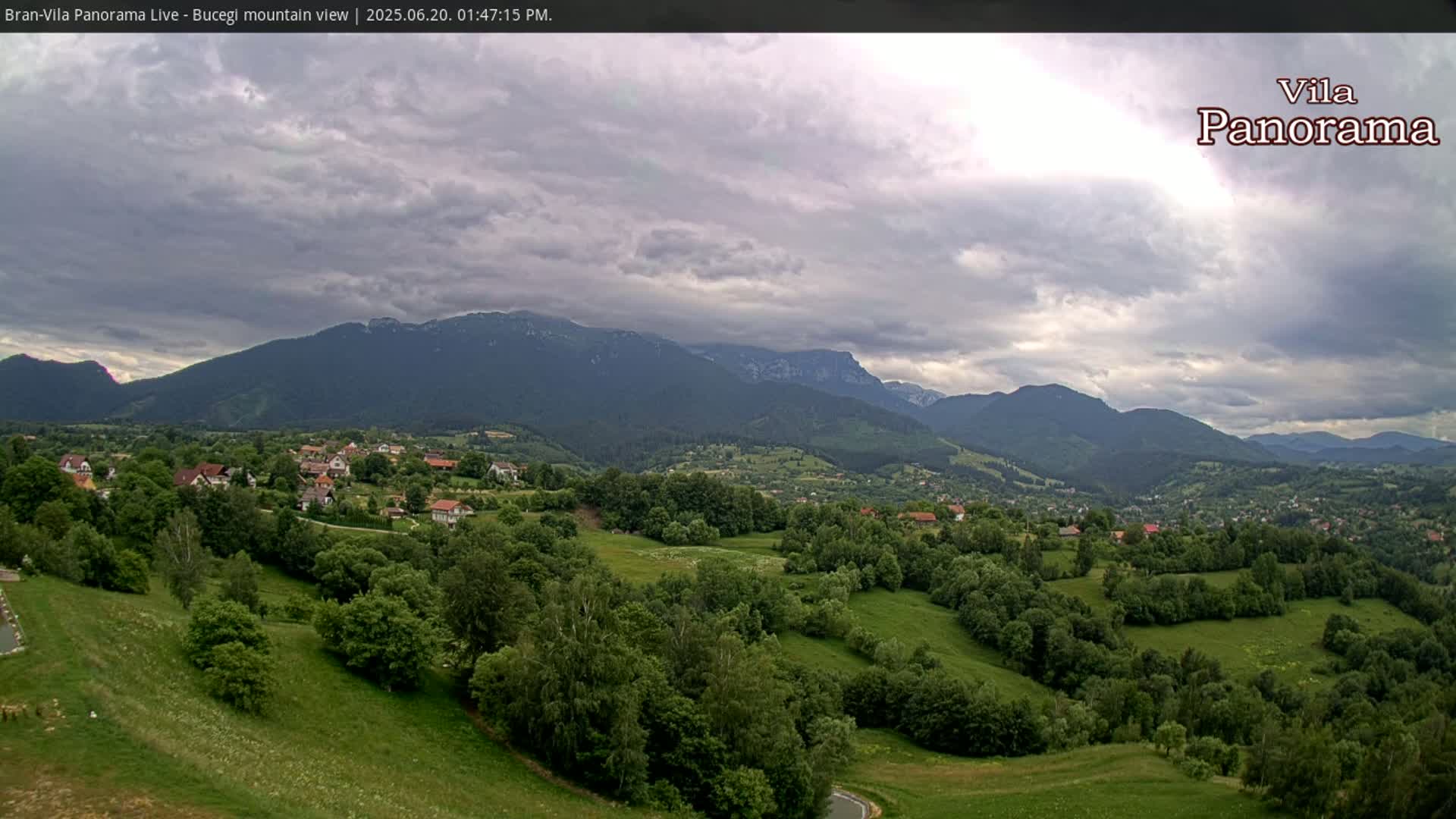 A lush green valley dotted with houses and trees stretches out beneath a cloudy sky, with imposing mountains in the background.