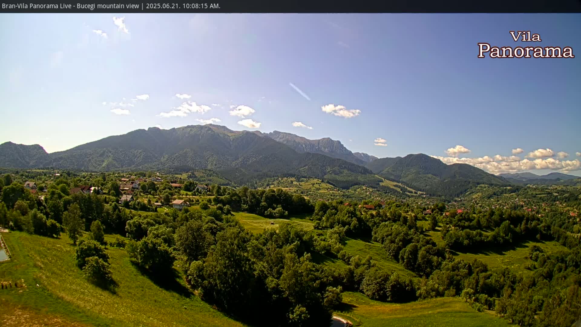 A sprawling green valley dotted with houses and trees, nestled beneath a range of mountains under a mostly clear blue sky with a few scattered clouds.