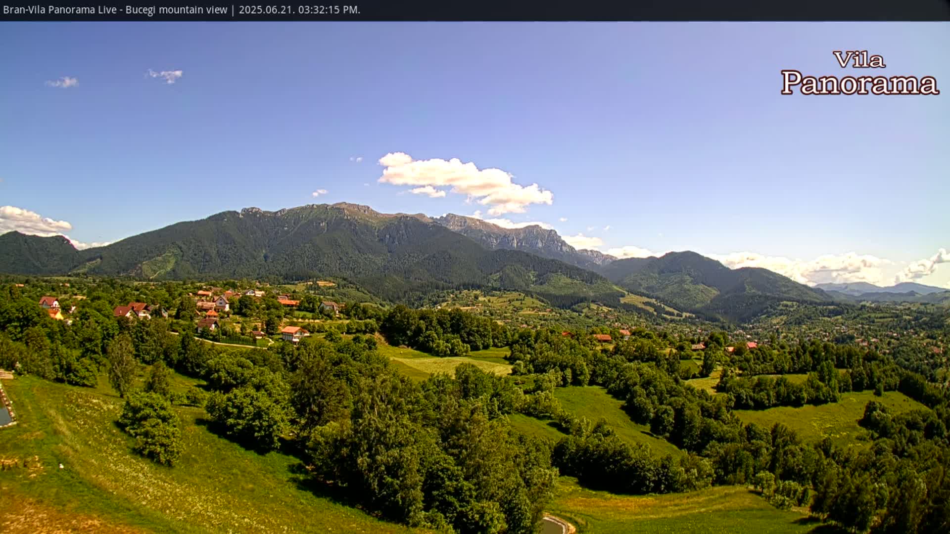 A verdant valley with a scattering of houses nestles beneath a range of mountains under a mostly clear, sunny sky.