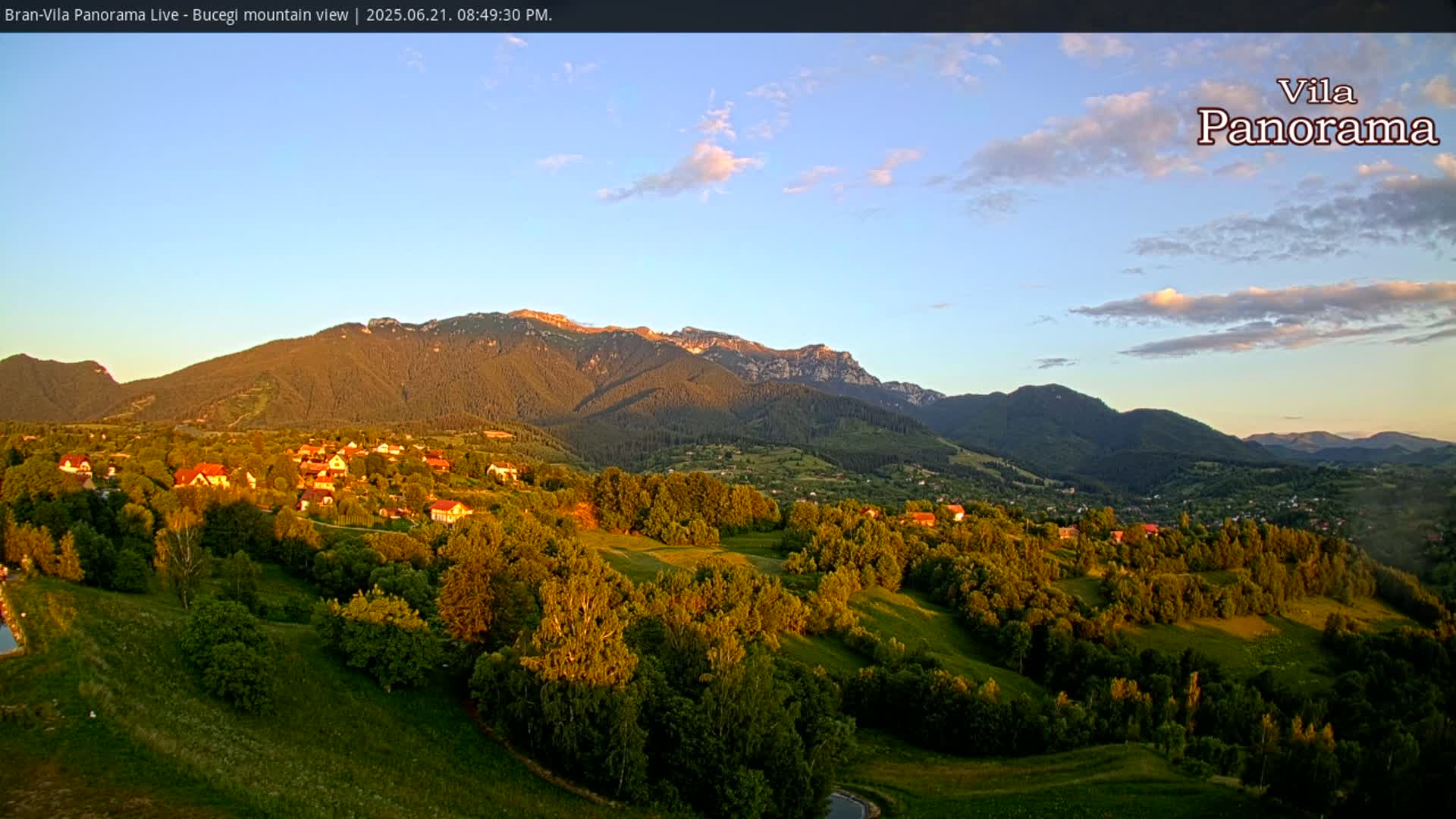 A sun-drenched village nestled in a valley, surrounded by lush green hills and mountains under a mostly clear, twilight sky.