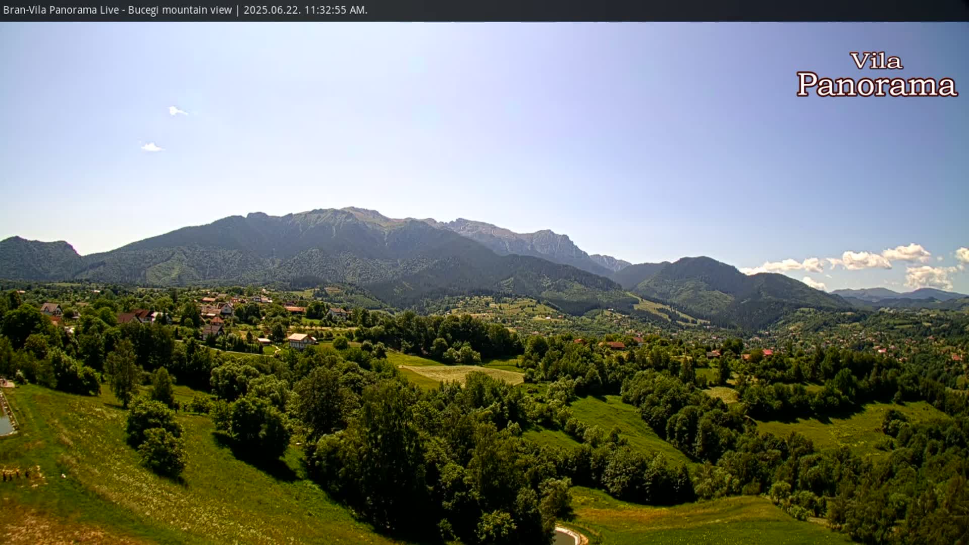 A sunny, clear day reveals a sprawling green valley dotted with houses and trees, nestled beneath a range of majestic mountains.