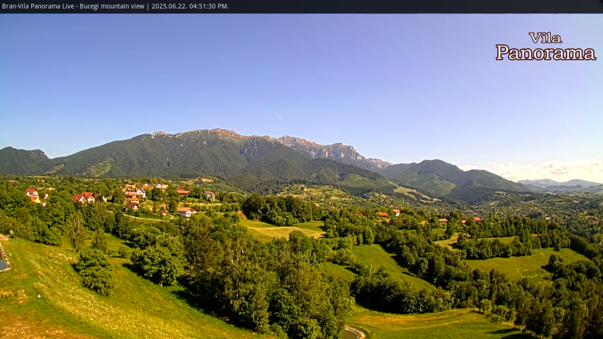 A small village nestled in a verdant valley, with a clear blue sky and a mountain range in the background under sunny conditions.