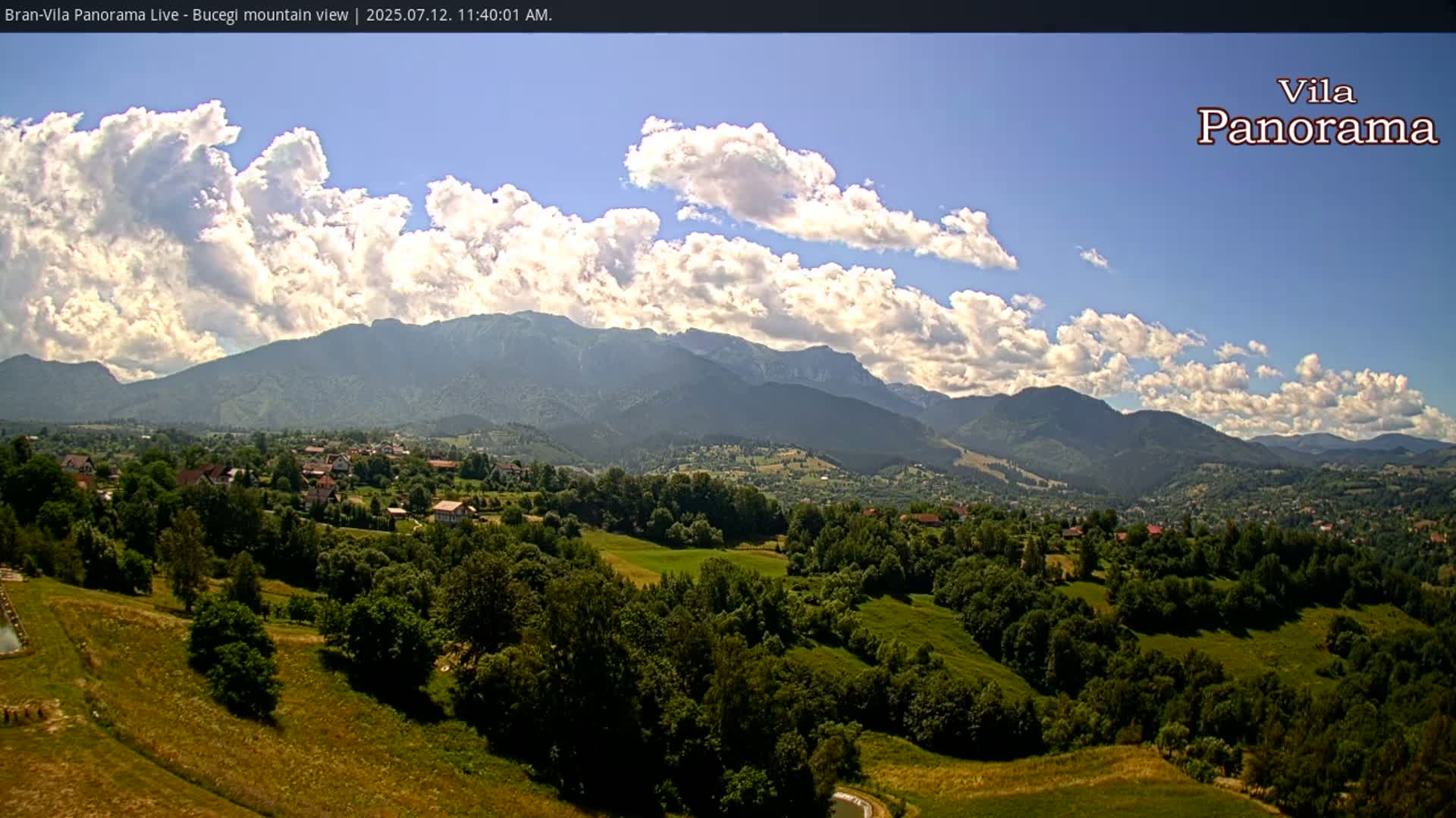A partly cloudy sky overlooks a lush green valley dotted with houses and trees, with a mountain range in the background.