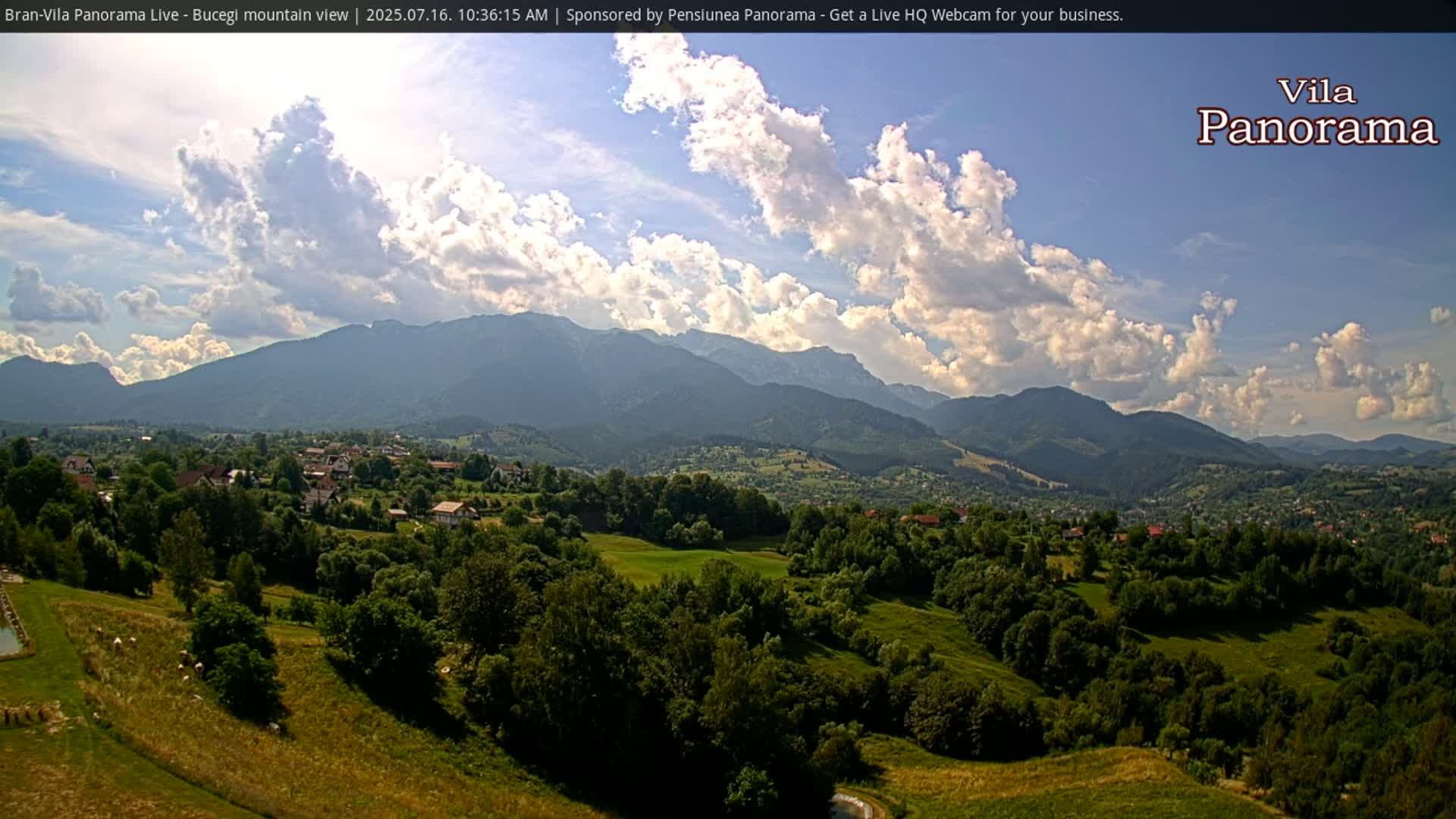 A partly cloudy sky overlooks a verdant valley dotted with houses and trees, nestled beneath a range of mountains.
