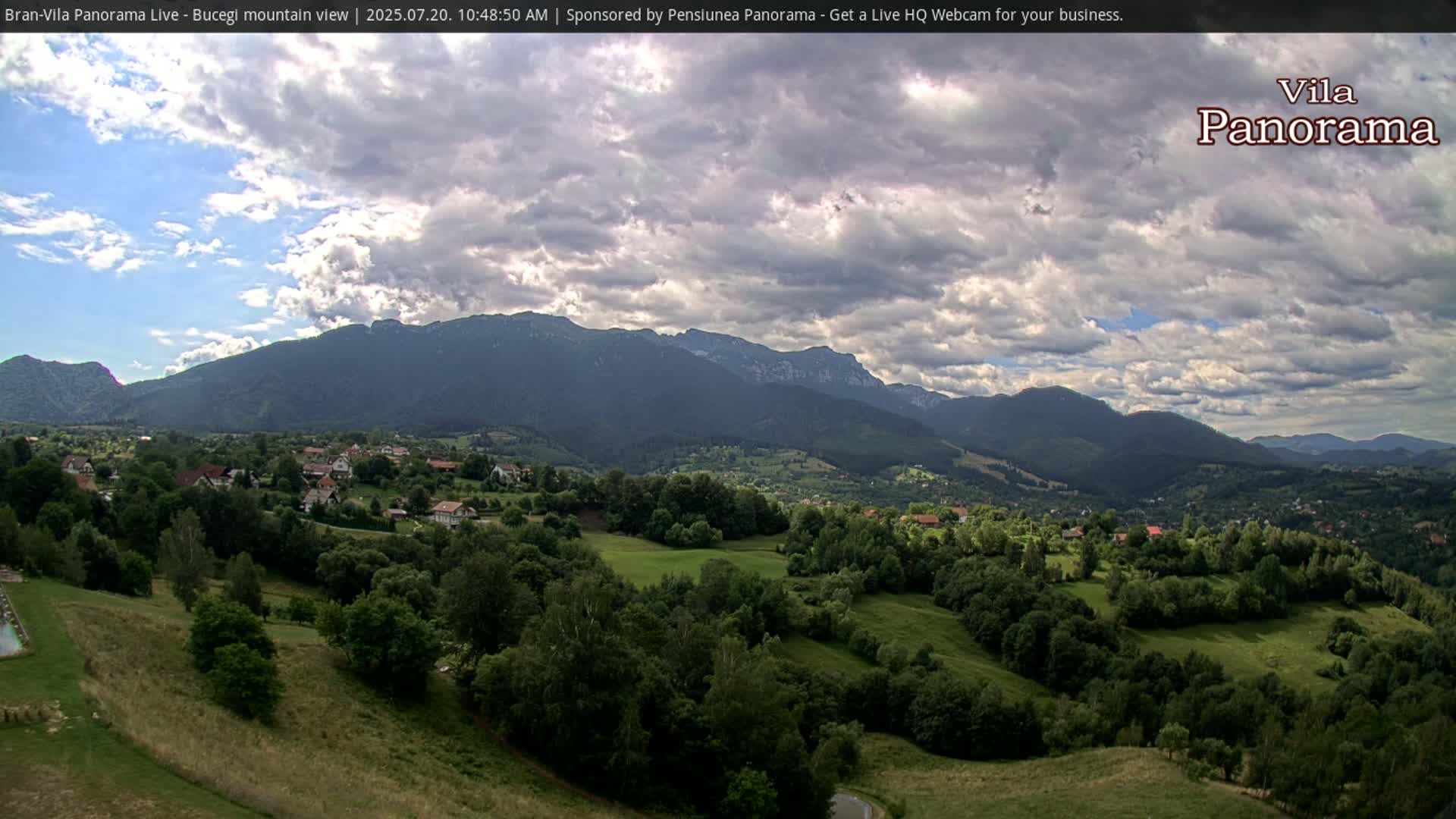 A partly cloudy sky overlooks a verdant valley dotted with houses and trees, nestled at the foot of a large mountain range.
