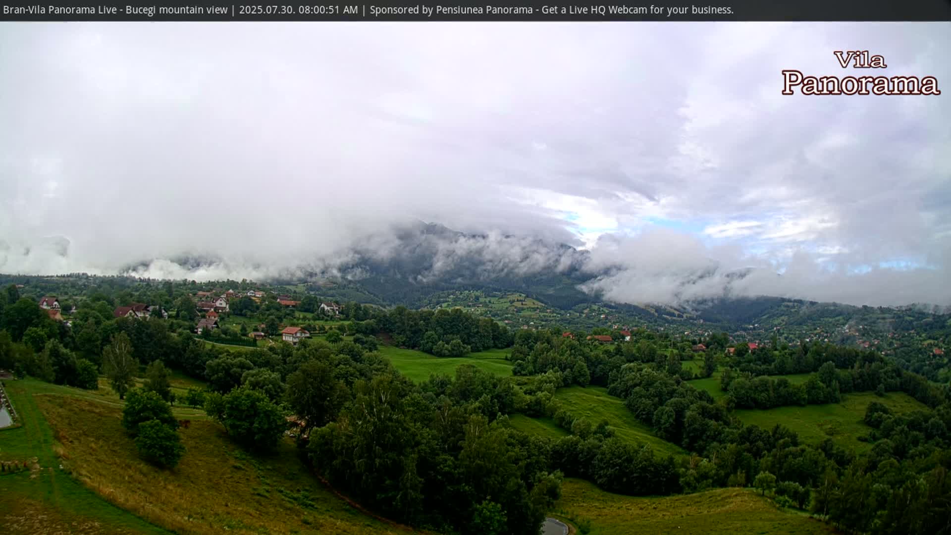 A partly cloudy sky overlooks a verdant valley dotted with houses and trees, nestled at the foot of a large mountain range.