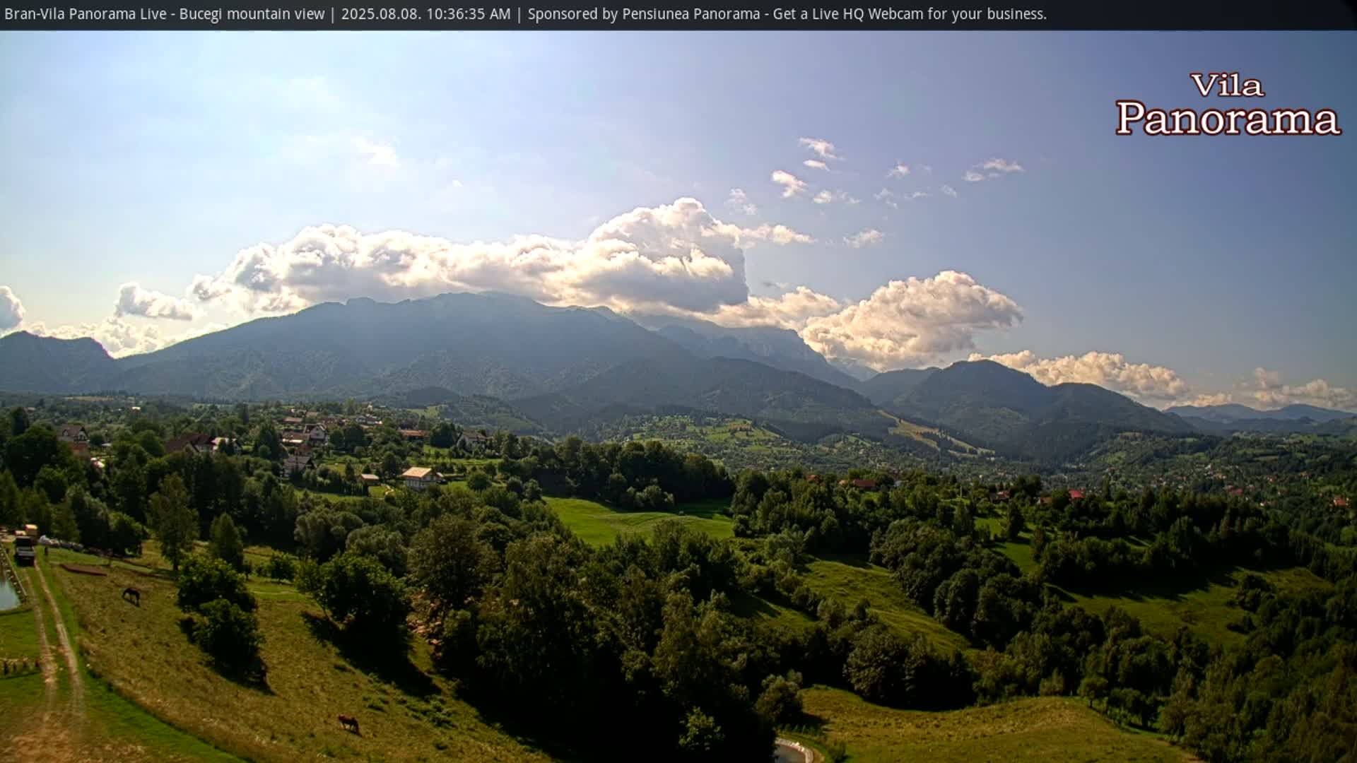 A partly cloudy day reveals a view of a village nestled in a green valley, with a mountain range in the background.