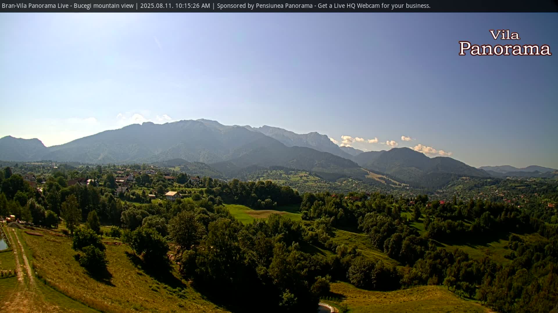 A sunny, clear day reveals a verdant valley dotted with houses and trees, nestled beneath a range of majestic mountains.