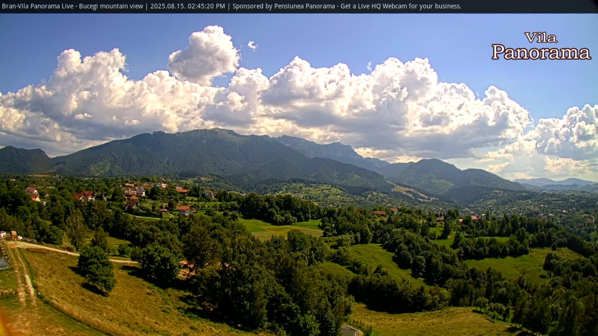 A sunny, clear day reveals a verdant valley dotted with houses and trees, nestled beneath a range of majestic mountains.