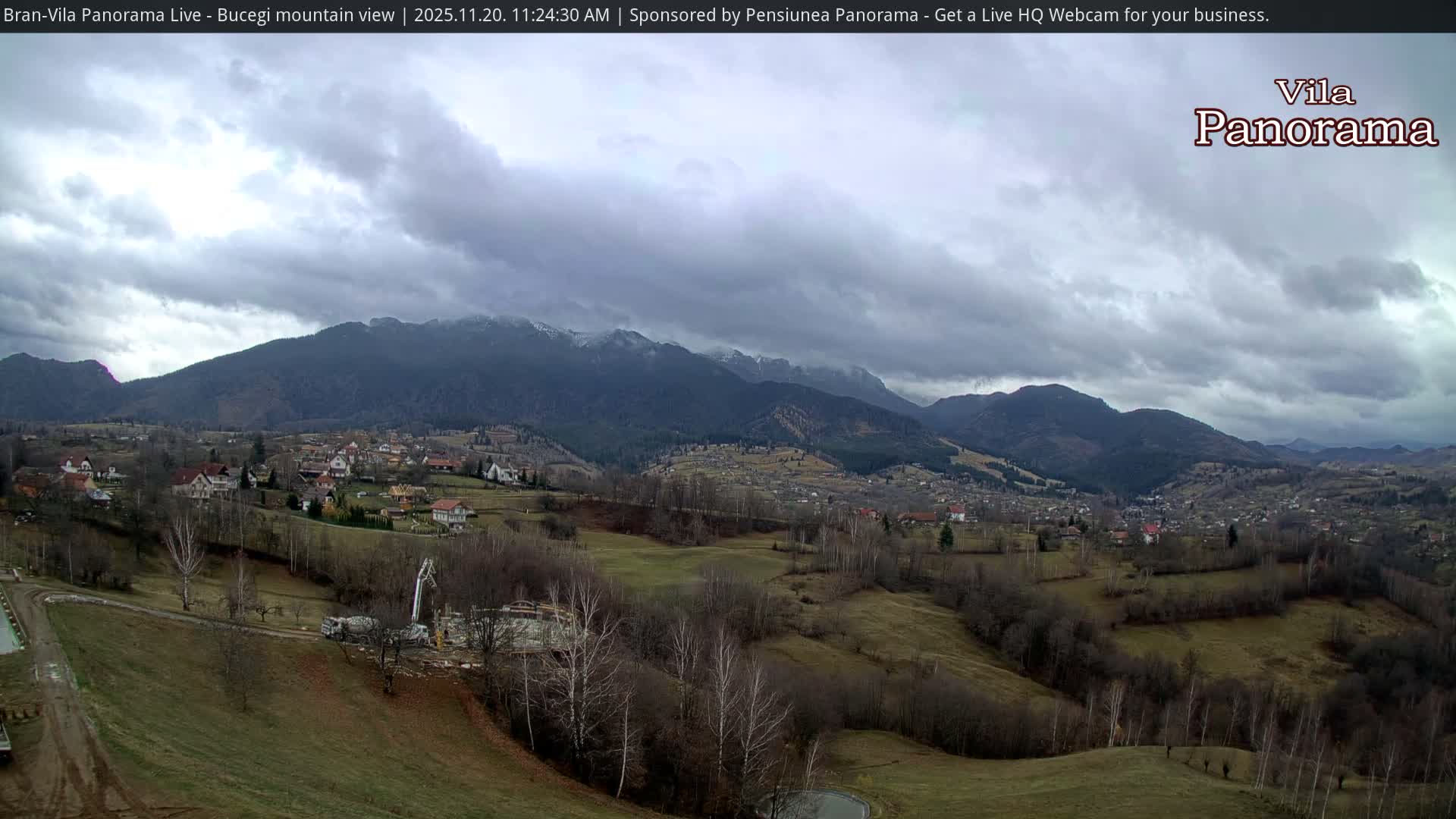 A wide panoramic view reveals villages nestled among rolling hills with dry, sparse vegetation and bare trees, backed by imposing, partly snow-capped mountains under a heavily overcast sky.