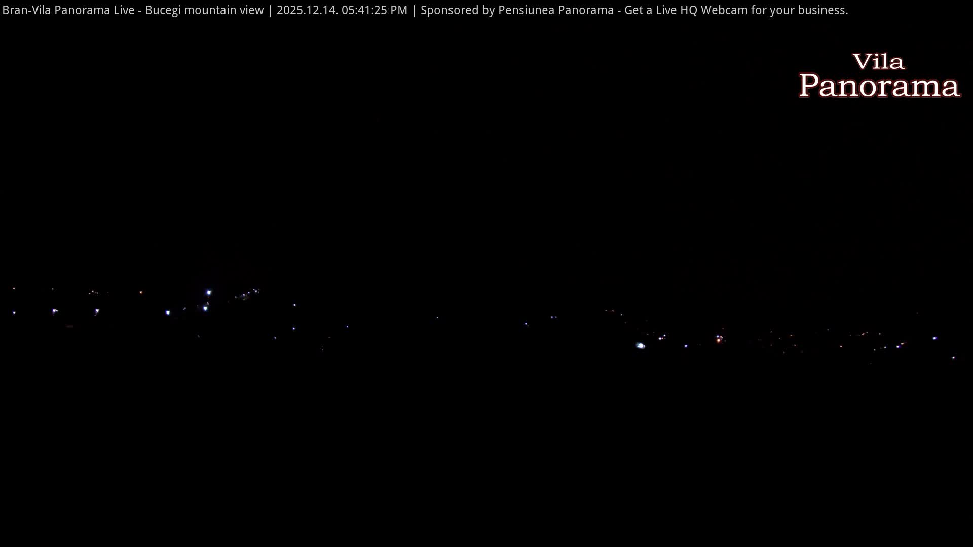 A wide panoramic view reveals a rural village nestled among rolling hills and bare trees, backed by majestic snow-capped mountains under a partly cloudy sky with bright sunshine.